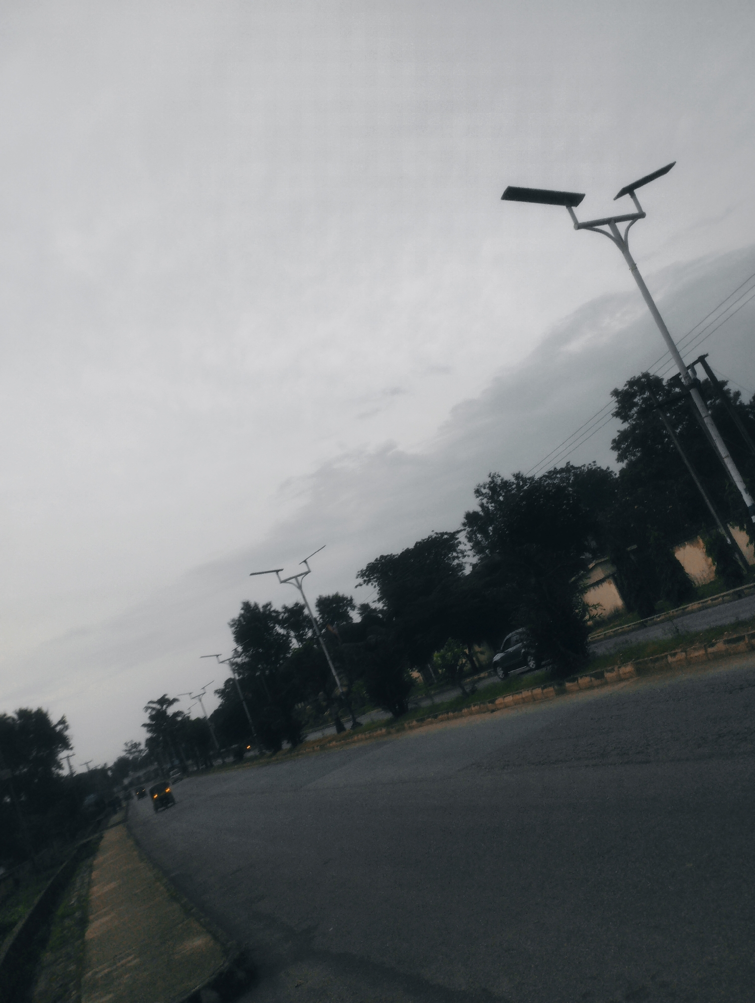 The image depicts an overcast sky over a street in Jos, Nigeria. The road, paved with asphalt, stretches diagonally across the frame. Along the left side of the road is a grassy verge with a concrete drainage channel. On the right side of the road, there is a yellow-striped curb, followed by a line of trees. Several lampposts with solar panels are visible along the road, some with two lights. A few vehicles are present, including a dark-colored car parked on the side of the road and another vehicle with its taillights illuminated driving away from the camera. The trees are dense and dark green, with some buildings partially visible behind them. The overall impression is that of a somewhat hazy, possibly humid, day in an urban or suburban environment.