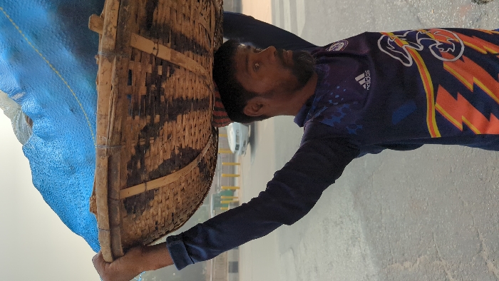 A male individual is observed in an outdoor urban setting in Dhaka, Bangladesh, carrying a large, circular, woven bamboo basket on his head. The basket's contents are obscured by a bright blue tarp, partially secured with yellow cordage. The man, with dark skin, a short beard, and dark hair, wears a long-sleeved dark blue athletic shirt featuring an Adidas logo, a white "3" emblem, and orange/yellow lightning bolt graphics. A small, striped cap is visible beneath the basket on his head. His arms are raised, hands supporting the basket's rim. His gaze is directed towards the viewer's left. The background reveals a paved road with yellow bollards or barriers and a white vehicle. The lighting is bright and diffuse.