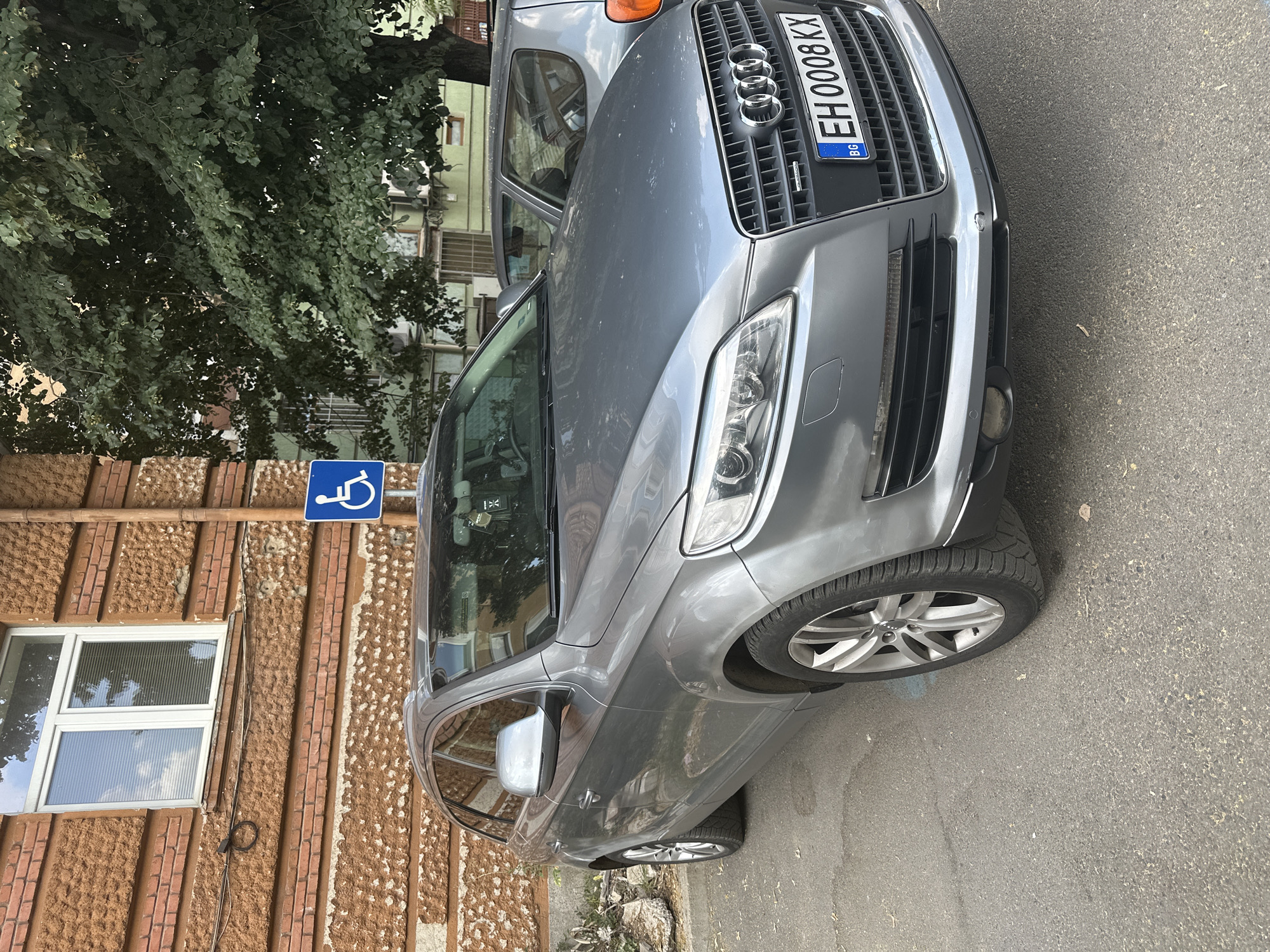 A grey Audi Q7 is parked on an asphalt surface in front of a brick building with a window. To the left of the car, there are some rocks and overgrown vegetation. A blue handicap parking sign is mounted on a pole to the right of the car. The car's license plate reads "EH 0008 KX" and has "BG" on the left side. Another car, a silver hatchback, is parked behind the Audi. A large tree with green leaves is located behind the cars and to the right of the building. The sky is visible through the leaves of the tree, reflecting in the window of the building. The building appears to be an older structure, with textured brickwork and a single, white-framed window. The overall scene suggests an urban environment, possibly a residential area or a public parking lot.