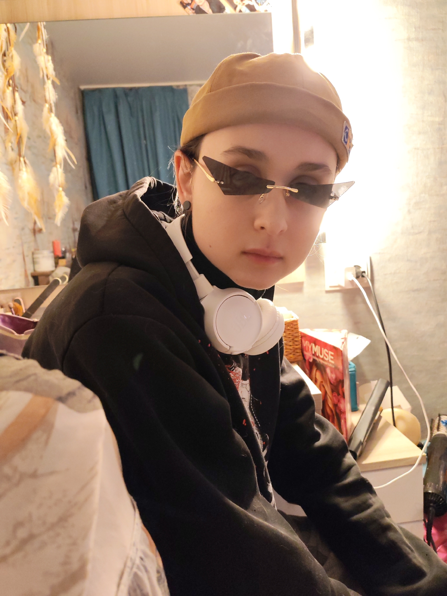 The image shows a young individual indoors in Chaikovsky, Russia. The person, positioned slightly left of center, is looking directly at the camera with a neutral expression. They wear a tan-colored beanie, dark-tinted futuristic sunglasses with gold-colored frames, a black hoodie, and a visible white turtleneck or collar beneath. White over-ear headphones rest around their neck. A black stud earring is visible in their left ear.
The background on the left side features a mirror reflecting a blue curtain or fabric. A decorative wall hanging with multiple light-colored feathers hangs against a textured, light-colored wall. On the right side, a wooden shelf is visible, holding a woven basket and a magazine titled "MYMUSE" featuring a person on its cover. A dark-colored electronic device, possibly a phone or tablet, leans on the shelf. An electrical outlet with white and black cables plugged in is mounted on the wall. A partially visible dark object resembling a hair dryer is situated to the far right. The scene is illuminated by an unseen light source from the right, creating some glare.