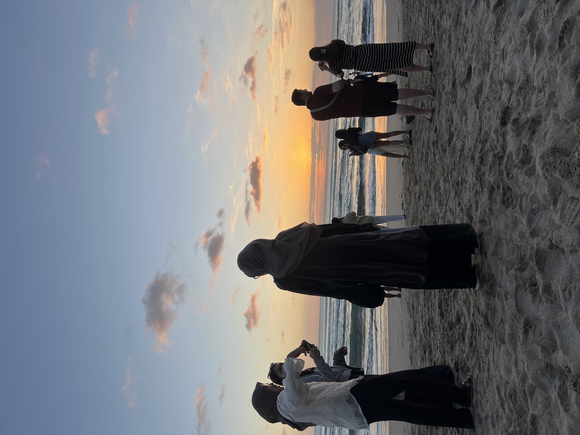 A group of people are standing on the sandy beach of Kuta, Bali, Indonesia, watching the sunset over the ocean. The sky is filled with warm hues of orange and pink from the setting sun, with scattered clouds reflecting the colors. The ocean waves are gently rolling onto the shore. Several individuals are taking photos with their phones, capturing the scenic view. The people are dressed in casual beach attire. The sand is coarse and shows footprints from previous visitors. The overall atmosphere is peaceful and serene, with a sense of awe and appreciation for the natural beauty of the sunset.