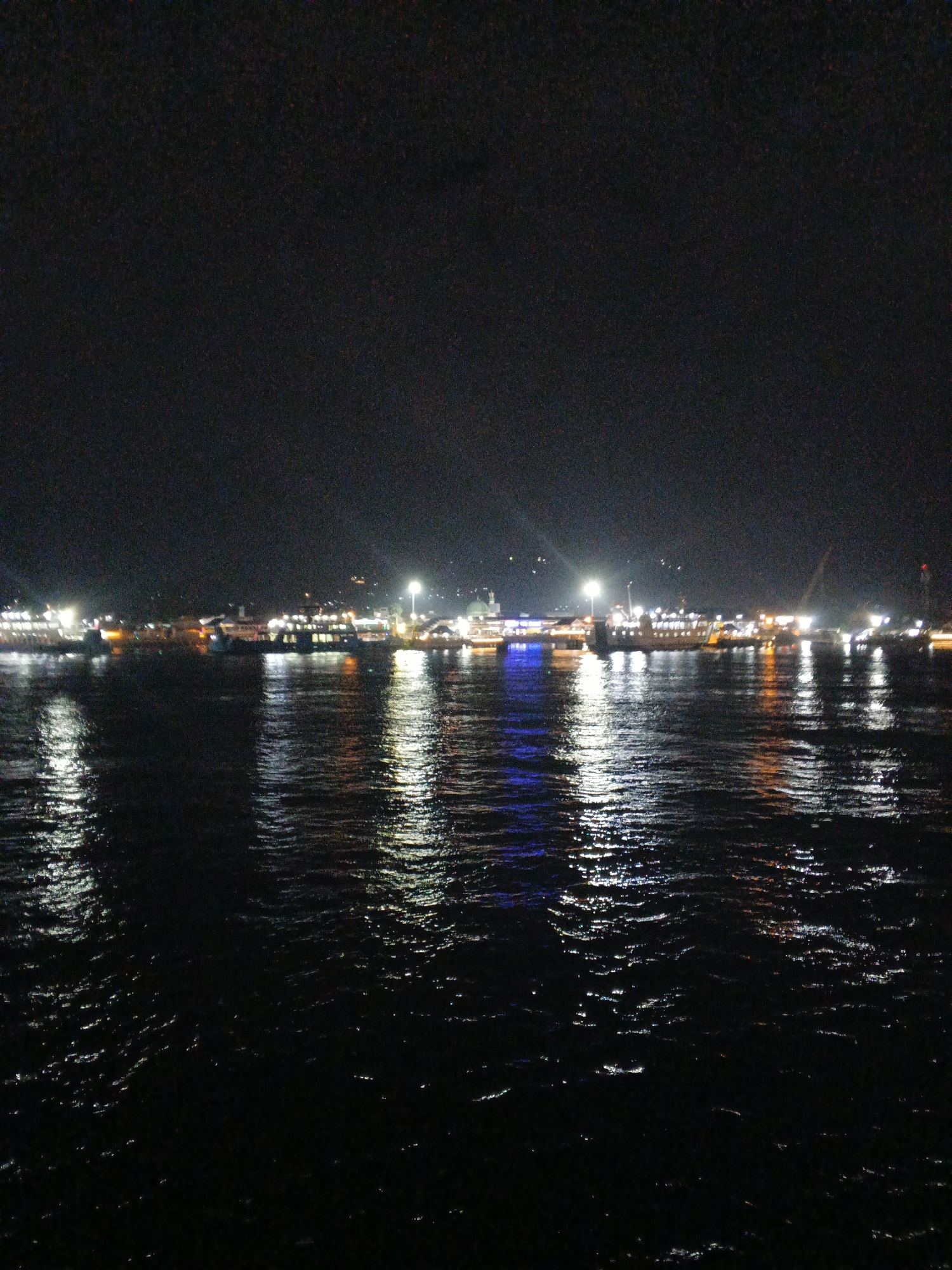 The image depicts a night scene with a body of water in the foreground and a city skyline with docked ferries in the background. The water reflects the lights from the city and ferries, creating a shimmering effect. The sky is dark, indicating it is nighttime. The city appears to be Banyuwangi, Indonesia, a port city. The ferries are illuminated, suggesting they are either docked for the night or preparing for departures. There are no people or specific actions visible in the image. The environment is characterized by the contrast between the dark night sky and the illuminated city lights reflecting on the water.