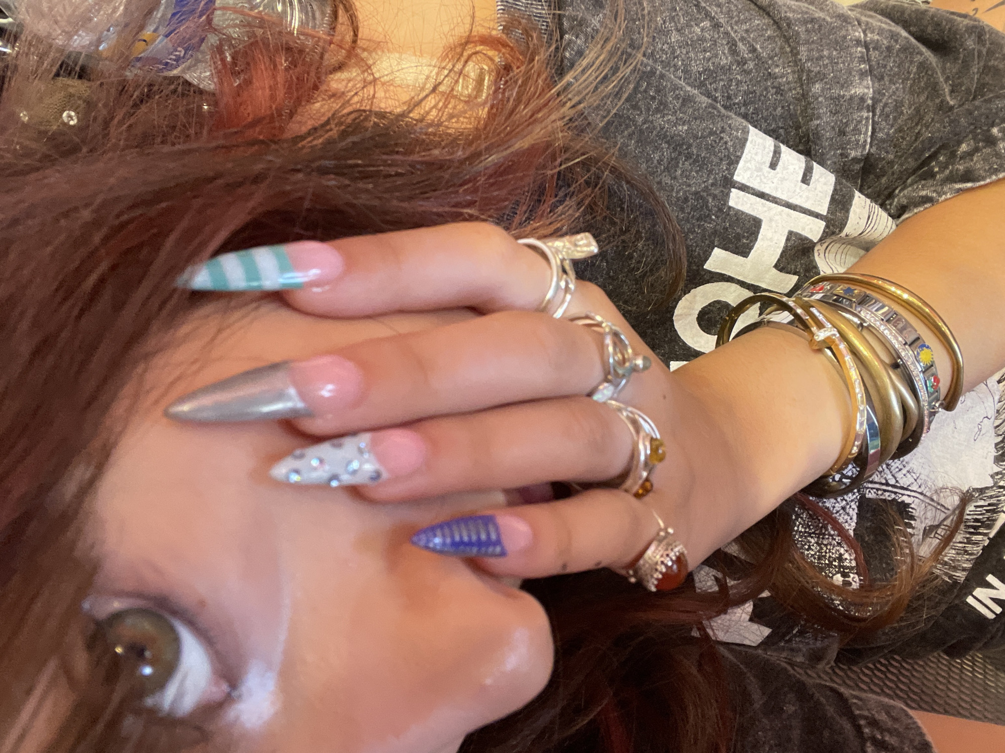 A close-up, eye-level shot shows a person's face and hand, adorned with elaborate fingernails and jewelry. The person has fair skin and reddish-brown hair, with visible makeup including white eyeliner or cream eyeshadow on the lower eyelid. The fingernails are long and pointed, decorated with various designs: one is silver, another has rhinestones and pink polish, a third is blue with metallic stripes, and a fourth features turquoise and white stripes. The hand is decorated with several rings, including silver bands and one with a reddish-brown stone, and multiple stacked bracelets, some appearing to be gold-toned and others with colored accents. The person is wearing a dark grey t-shirt with white text that is partially visible. The background is softly focused and appears to be indoors, with hints of warm lighting. There is no indication of the city of Raanana, Israel, in this image.
