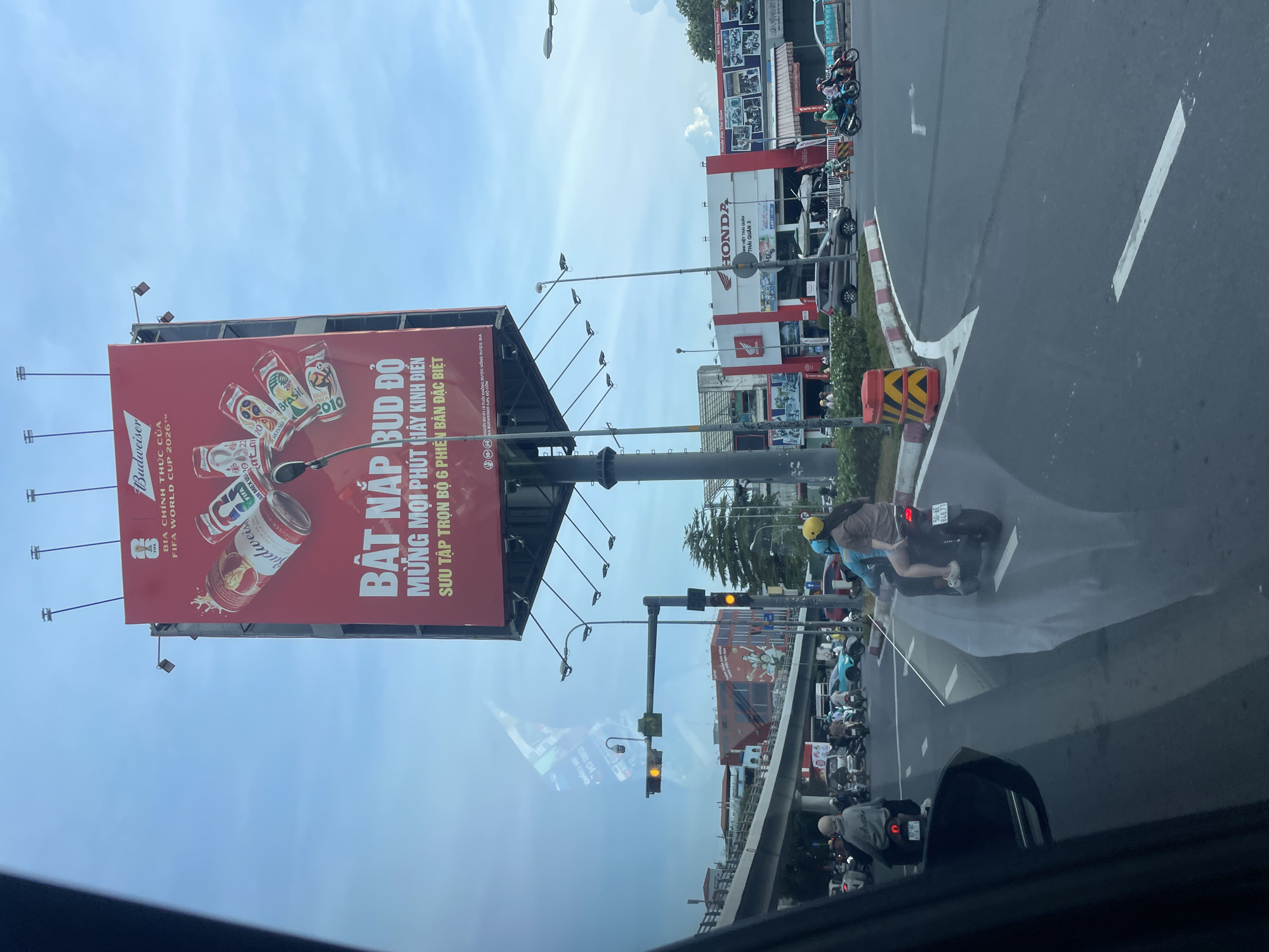 This is a photograph of a street scene in Thủ Đức, Vietnam, taken from inside a vehicle, likely a car, given the perspective through the windshield and the visible side mirror. The primary subject is a large red billboard advertising Budweiser beer, prominently featuring the FIFA World Cup 2026™ logo and imagery of beer cans. The billboard is mounted on a tall pole structure.
In the foreground, the road is visible with traffic lines and several motorcycles. Two individuals are riding on a motorcycle in the right lane, the passenger wearing a yellow helmet and a brown shirt, and the driver wearing a blue shirt. Other motorcycles and vehicles are present on the road.
To the right of the road, a Honda dealership is visible, with the "HONDA" logo clearly displayed. Several motorcycles are parked in front of the dealership.
The environment appears to be an urban setting with traffic lights, streetlights, and overhead infrastructure, likely an overpass. The sky is mostly clear with some clouds. The reflection on the windshield of the vehicle reveals some of the surrounding environment, including buildings and possibly other advertisements. The overall lighting suggests it is daytime.