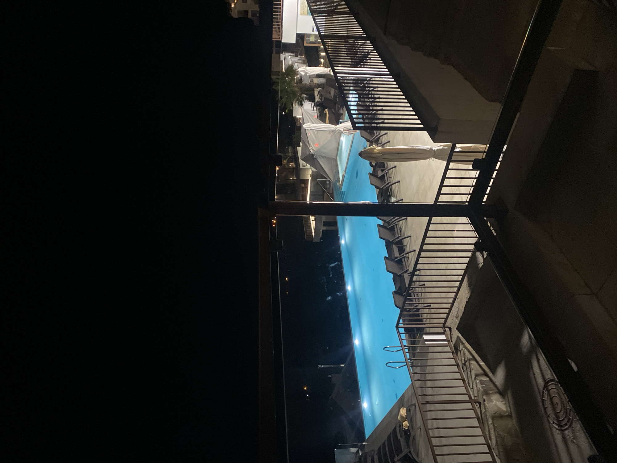 This is a nighttime, high-angle shot of an illuminated swimming pool area, likely at a resort or hotel, in Uch-Dere, Russia. The pool is a bright, turquoise blue and is lit from beneath. Several lounge chairs are arranged around the pool, some occupied by two dogs resting. A metal railing separates the viewer from the pool area. In the background, there are structures and palm trees, and the lights of a distant town are visible. The overall atmosphere is serene and tranquil, with the dark sky contrasting sharply with the vibrant blue of the pool.