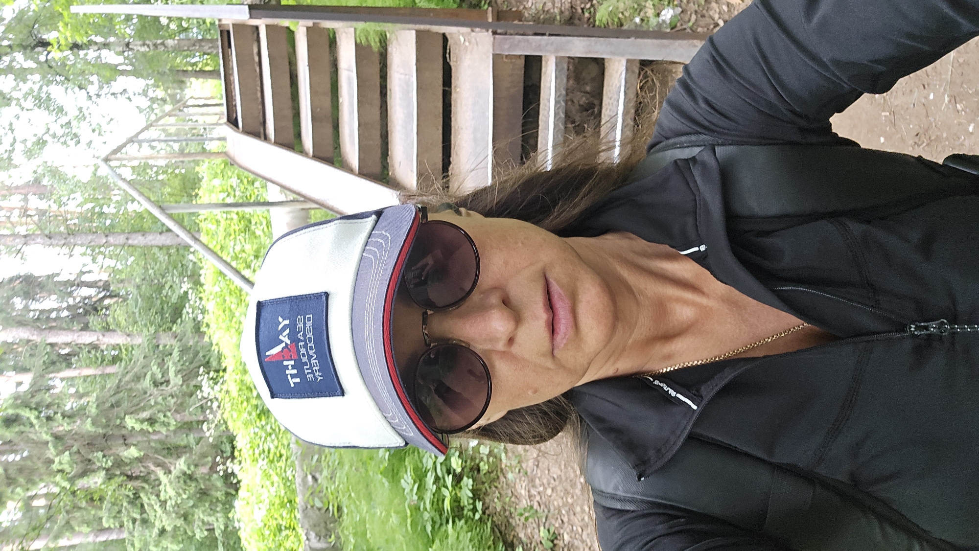 A woman wearing a black zip-up jacket, sunglasses, and a white and gray baseball cap with a logo stands in front of a metal staircase in a wooded area. The cap has a dark blue patch with white text that appears to be reversed, reading "TH AY" at the top and "SEA ROUTE DISCOVERY" below. The staircase has metal steps and railings and ascends to the right of the frame. The background consists of trees, greenery, and a dirt path. The image appears to have been taken outdoors during daylight. The woman is looking directly at the camera. The location is identified as the city of Chaikovsky, Russia.