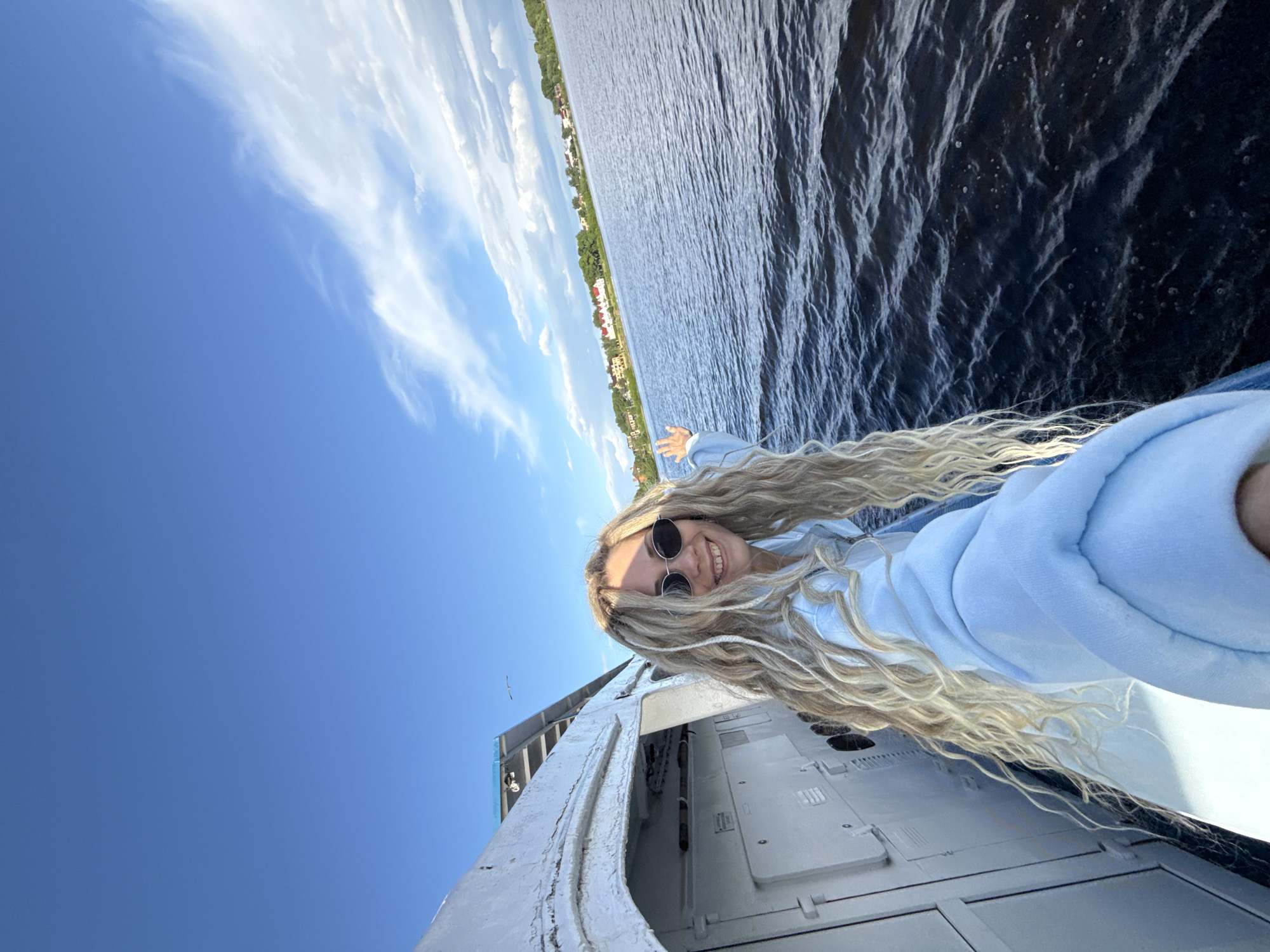 A selfie depicts a woman with long, blonde, wavy hair, wearing round sunglasses and a light blue hooded sweatshirt, standing on a gray and white structure, possibly a boat or building, with the sky and water visible in the background. The picture is taken at an angle, making the landscape appear tilted. The sky is a bright blue with wispy white clouds. The body of water is dark, with some visible waves. The cityscape of Заволжье, Russia is visible in the distance across the water.