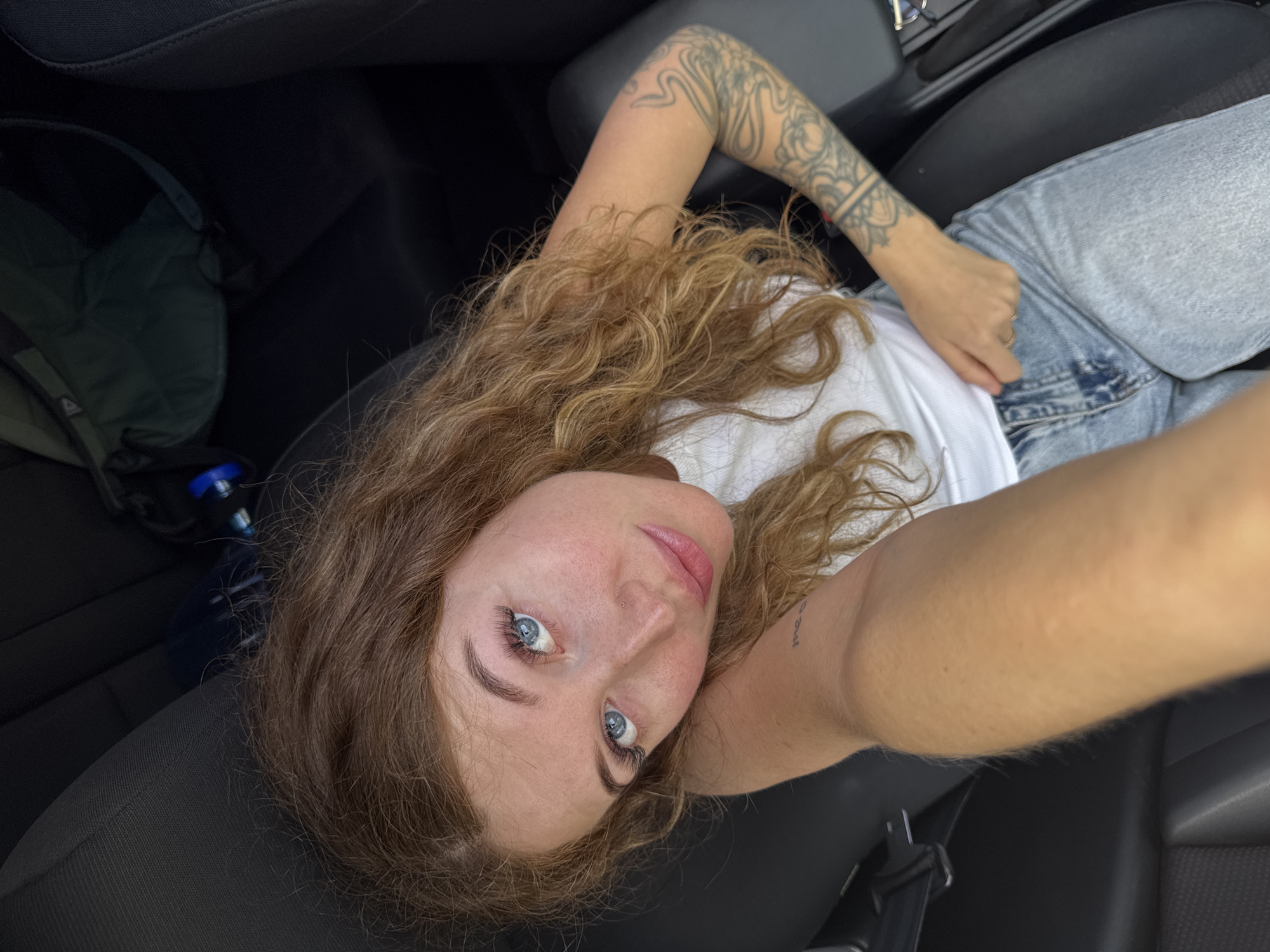A young woman with light brown, curly hair and blue eyes is taking a selfie inside a vehicle. She is wearing a white t-shirt and light blue jeans. Her left arm has a tattoo. A black backpack is on the back seat. The interior of the vehicle is dark gray. The photograph may have been taken in Chaikovsky, Russia but there is no evidence in the image to confirm that statement.