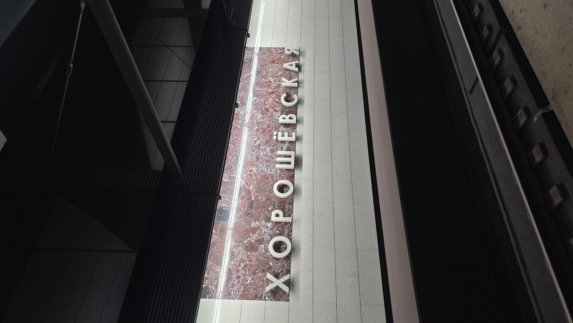 The image depicts the interior of a Moscow Metro station. The station name "ХОРОШЕВСКАЯ" (Khoroshyovskaya) is prominently displayed in large, three-dimensional white letters against a backdrop of red marble-patterned tiles and white stone tiles. The station features a dark ceiling with visible structural elements and lighting. In the foreground, a section of train tracks with ballast and ties is visible, indicating the active train platform. The overall impression is one of a modern, well-maintained subway environment.