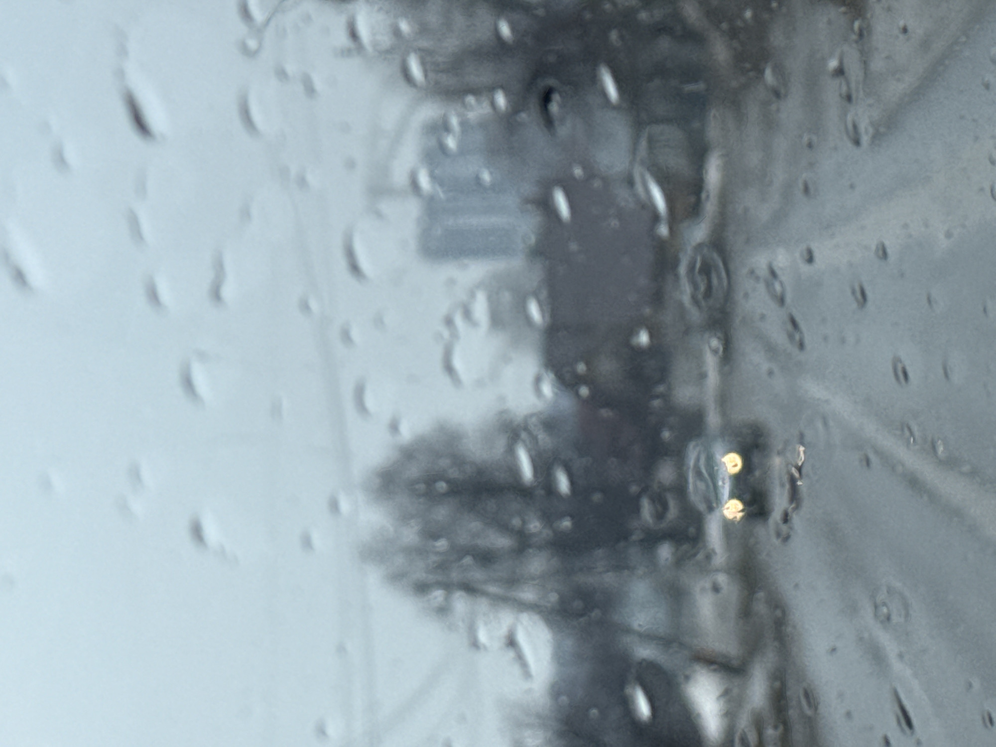 The image, likely taken in Chaikovsky, Russia, shows a view through a rain-streaked window. Outside, a street scene is visible with a vehicle, indicated by its headlights, moving in the distance. Trees and infrastructure, such as power lines or traffic signals, are also discernible, though blurred by the rain and distance. The general environment suggests a cloudy, rainy day.