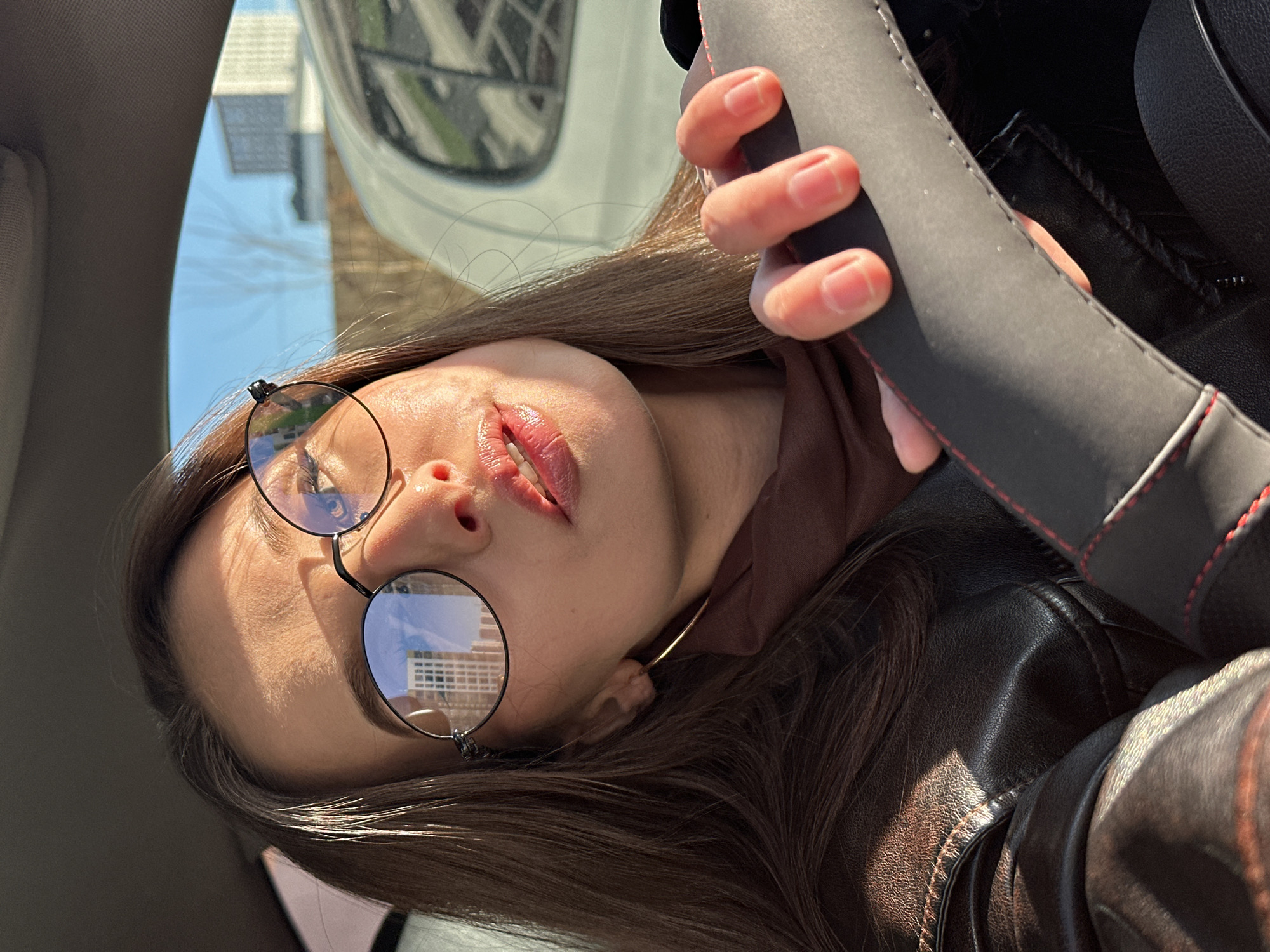 A young woman is captured in a close-up, diagonally oriented image taken inside a car. She has long, straight dark brown hair, which falls across her shoulders. She wears round, wire-rimmed glasses that reflect the surrounding environment, showing grid-like patterns of windows from nearby buildings. Her lips are slightly parted, revealing some teeth, and appear glossy.
She is dressed in a dark brown leather jacket or coat with visible stitching, and a lighter brown garment is visible underneath. Her right hand (viewer's left) is firmly gripping a black steering wheel cover accented with red stitching. Strong, direct sunlight illuminates her face and hair, creating distinct highlights and shadows, suggesting a bright midday or early afternoon setting on a clear day.
Through the car's windshield, a sunny outdoor scene is visible. Structures resembling multi-story buildings with windows are reflected in her glasses and can also be seen more broadly through the glass. A building with a light facade and a peaked roof is discernible against a clear blue sky. Parts of the car's interior, including the headliner, are visible in the foreground. The scene is set in Yekaterinburg, Russia. The woman's expression is posed and serene. There is no visible text.