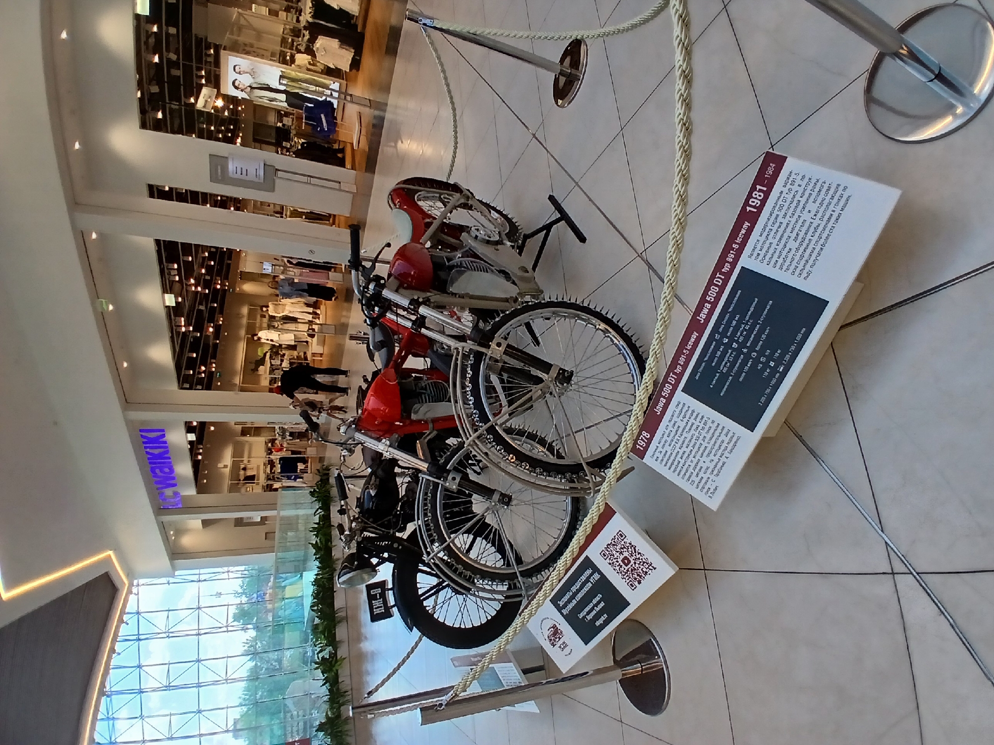 In Yekaterinburg, Russia, within a modern shopping mall with a large glass facade and a high ceiling featuring an illuminated ceiling design, two vintage motorcycles are displayed on a tiled floor, cordoned off by decorative ropes. One motorcycle is black, with a license plate reading "KM-B," and the other is red. Both have intricate spoke wheels. Informational plaques with QR codes are placed in front of the motorcycles. People are visible in the background, shopping in the stores, including one wearing a red hat. The stores in the background include "LC WAIKIKI."
