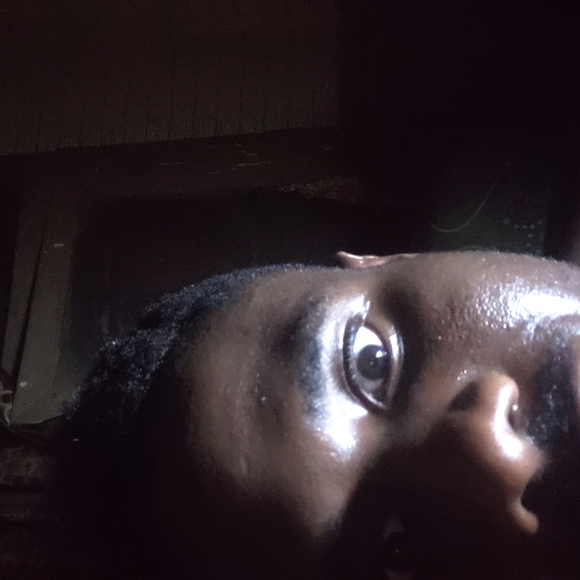 A close-up, low-angle photograph depicts a person's face, presumably in Narayi, Nigeria. The subject has dark skin, short dark hair, and a prominent eye visible to the viewer. The skin appears shiny, possibly due to sweat or oil. The background is dark and indistinct, suggesting a dimly lit or nighttime environment. A dark, textured surface is slightly visible in the upper left, possibly part of a wall or object. The composition focuses on the person's face, with the rest of the body and surroundings obscured.