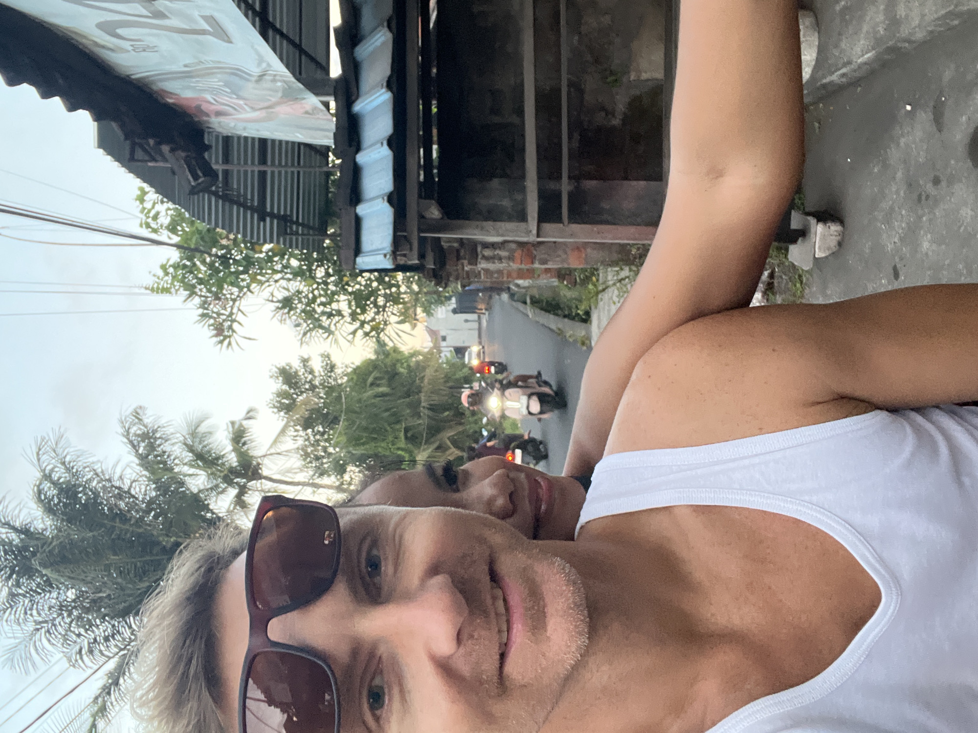 A selfie image captures a couple on a moped in the city of Sukawati, Indonesia. The man in the foreground wears sunglasses and a white tank top, smiling at the camera. Behind him, a woman is seen smiling as well. In the background, a street scene unfolds with other mopeds traveling in the same direction. The architecture includes buildings with corrugated metal roofs and brickwork, partially obscured by foliage. The sky is overcast, suggesting early morning or late afternoon lighting.