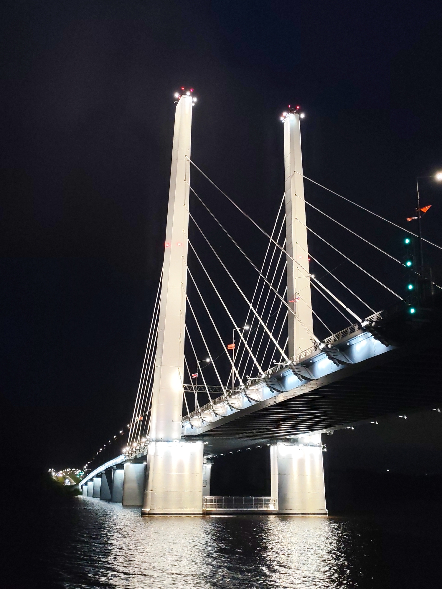Captured at night, the image reveals a cable-stayed bridge in the city of Unknown, Unknown, illuminated against the dark sky. The bridge features two prominent, white towers, each topped with bright lights. Cables extend from the towers to the bridge deck, which is lit with blue and white lights. The supporting piers of the bridge are also brightly illuminated. The water beneath the bridge reflects the lights, adding to the overall brightness of the scene. The environment is dark, with minimal visible detail beyond the bridge and its immediate surroundings.