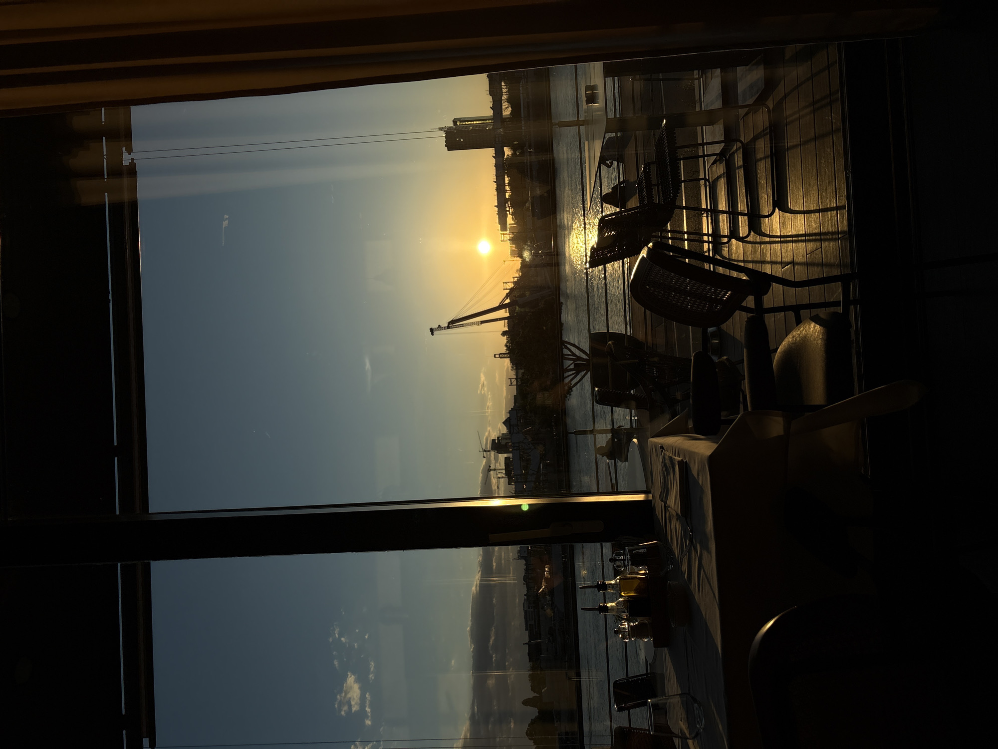 The image shows a sunset view through a window, likely from a restaurant in Belgrade, Serbia. The setting sun is visible over a body of water, reflecting light onto the surface. Silhouettes of buildings and cranes dot the skyline. The foreground includes tables and chairs, suggesting an indoor dining area. On one table, a set of oil and vinegar bottles is present. The scene has a warm, golden hue from the sunset.