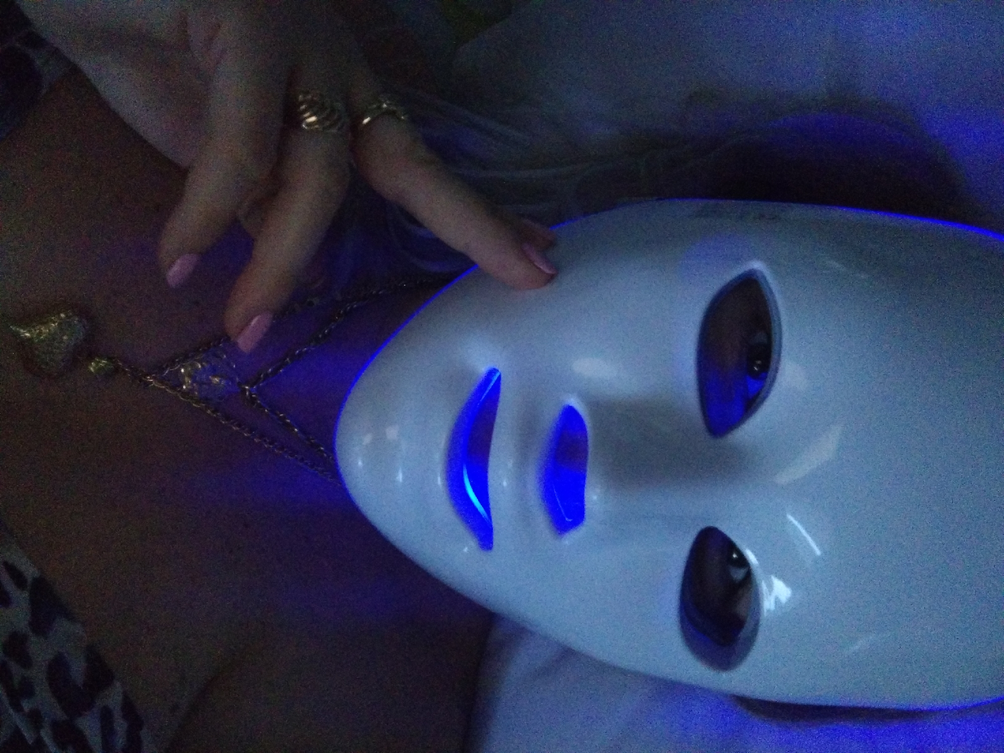 A close-up, over-the-shoulder shot shows a person wearing a white LED facial mask. The mask is illuminated with blue light, particularly around the eye and mouth openings. A person's left hand, adorned with rings, is visible near the mask. The person is wearing a gold necklace with a pendant. The background is dark with a hint of blue light. No specific location in Stara Zagora, Bulgaria can be determined from the image.