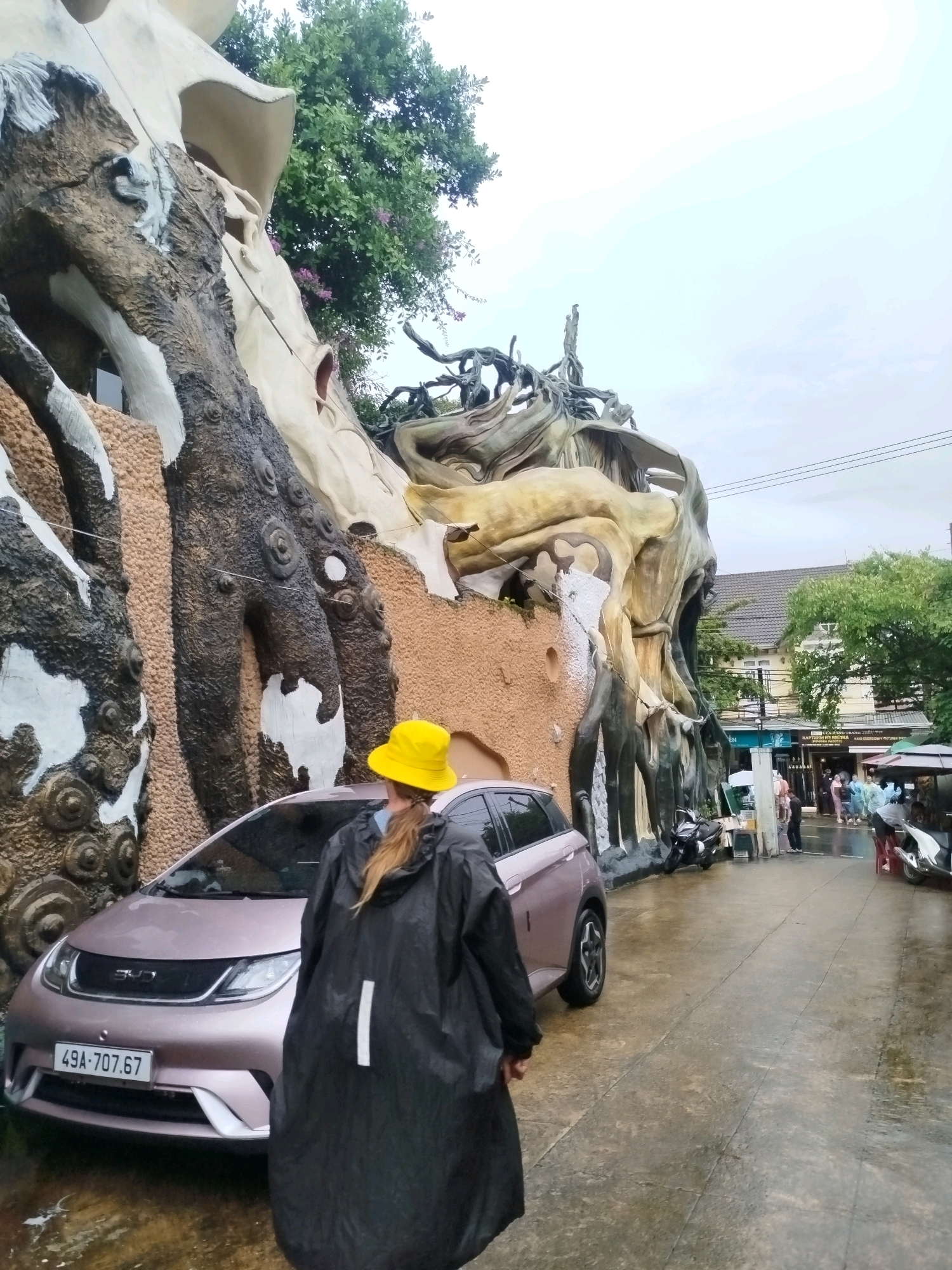 The image shows a woman wearing a black raincoat and a yellow hat standing in front of a light pink car. The car has a license plate that reads "49A-707.67". Behind the car is a large, abstract structure that resembles intertwined trees and roots. The ground is wet, suggesting it has recently rained. In the background, there are buildings, more trees, and other people. The architecture of the building in the foreground is highly unusual and organic. This is the Hang Nga Guesthouse, also known as the "Crazy House", in Da Lat, Vietnam, a surrealist architectural marvel designed by architect Dang Viet Nga.