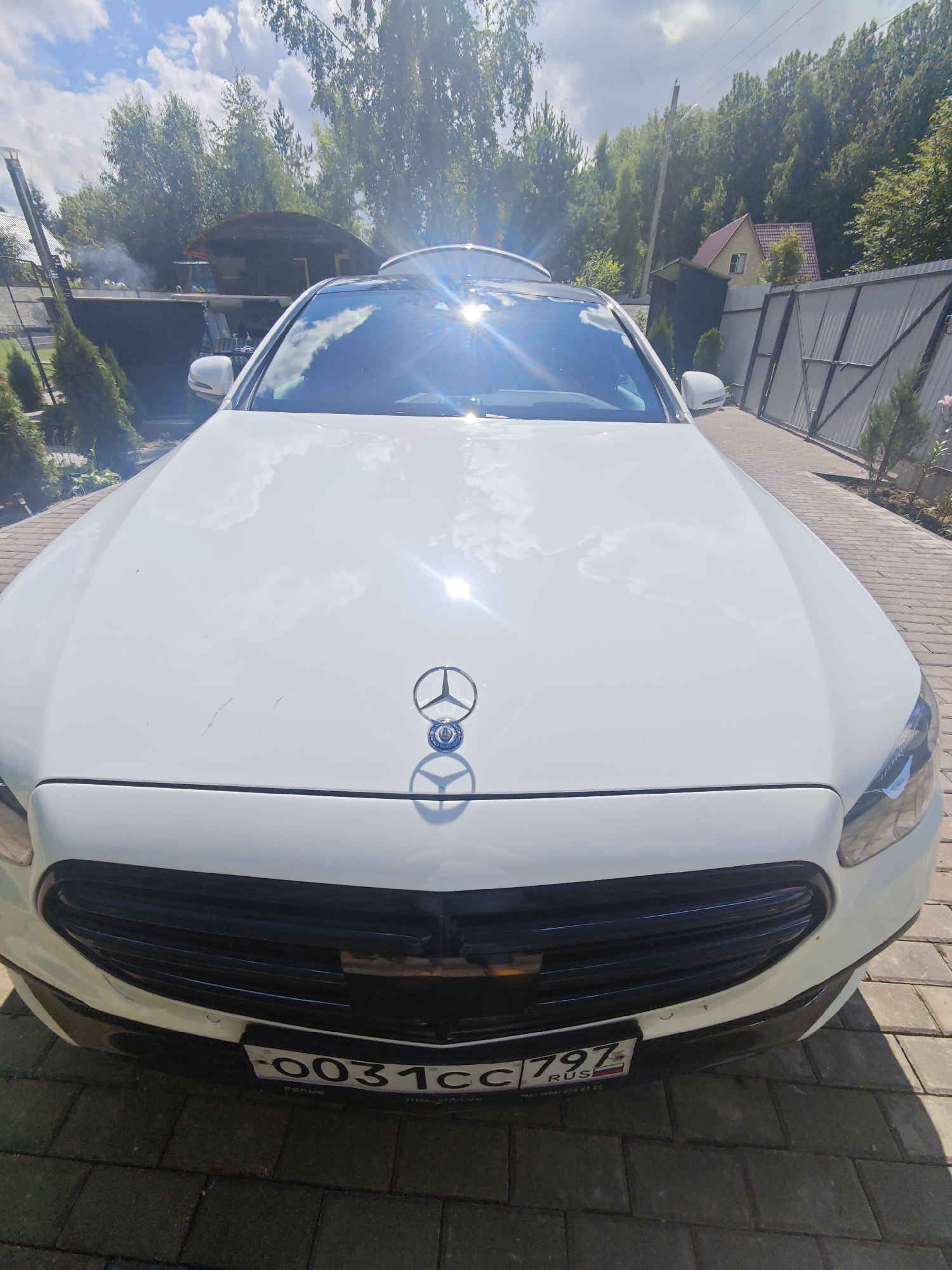 A white sedan is parked on a paved driveway in front of a house. The car has a Mercedes-Benz logo on the hood. The license plate is visible and reads "0031CC 797 RUS". In the background, there are trees, a fence, and a house with a red roof. The sky is blue with some clouds, and the sun is shining brightly, causing lens flare on the car's hood and windshield. The location is identified as Лужники Снт, Russia.