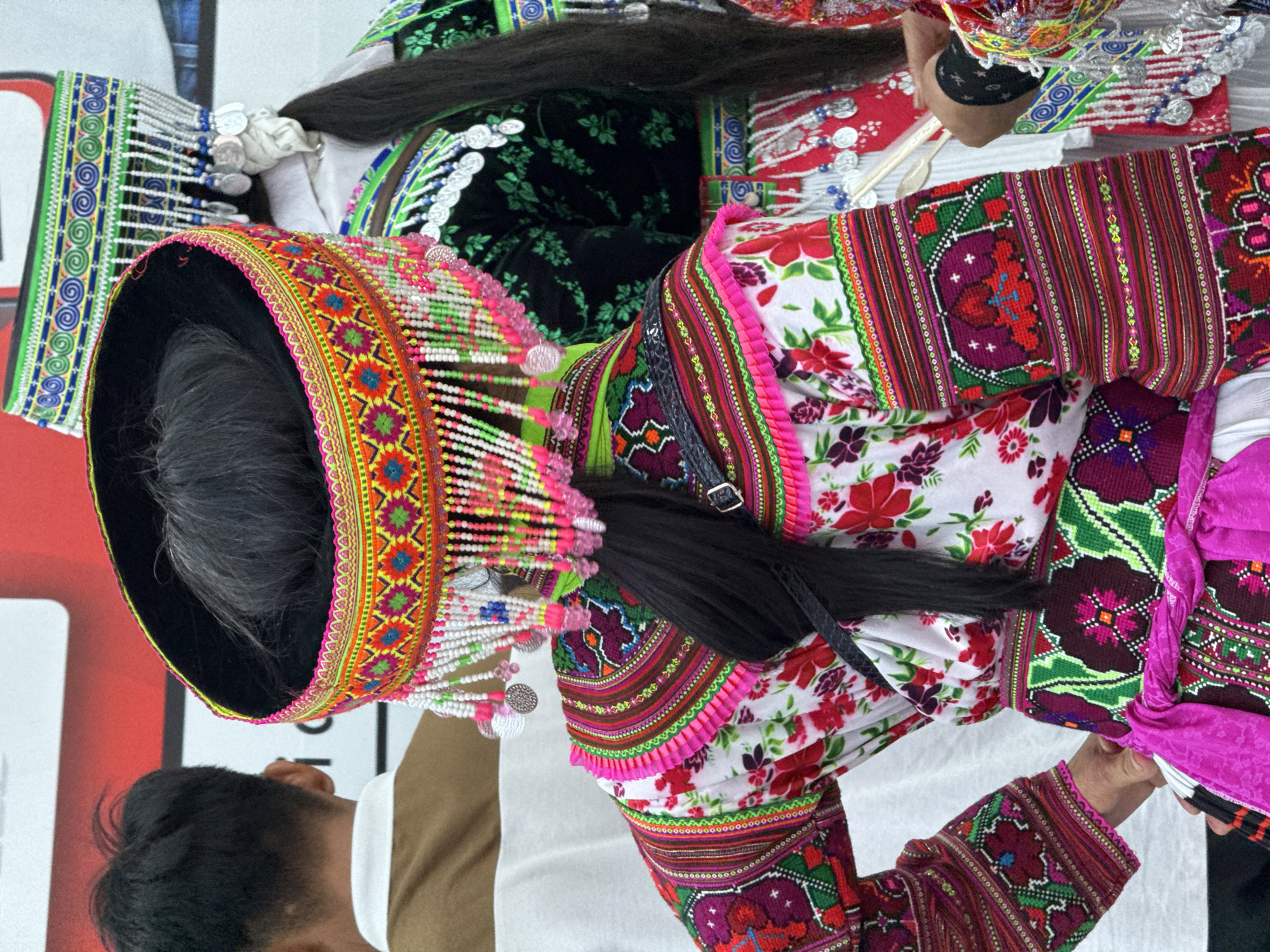 A vibrant image depicts a woman in a traditional Hmong outfit, likely a stage performer, standing in what is likely Da Lat, Vietnam. She wears a tall, ornate headdress adorned with multicolored beads, embroidery, and dangling silver coins. Her floral blouse has intricate embroidered details, and a decorative woven belt is visible. A person, likely an assistant or fellow performer, is seen partially in the lower left corner. The background suggests an indoor setting, perhaps a performance venue. The photograph focuses on the details of the costume.