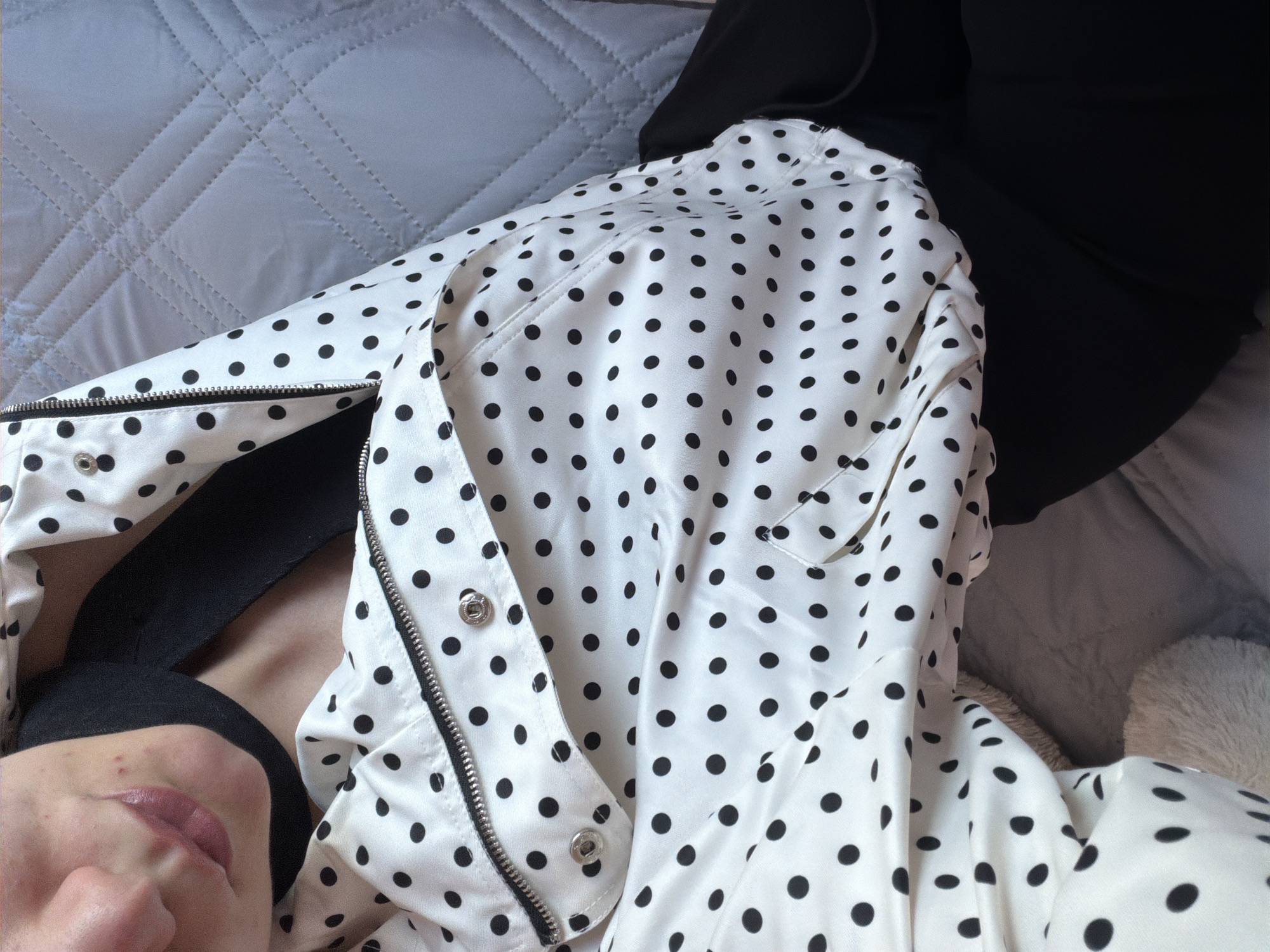 The image shows a close-up view from a low angle, suggesting the perspective of someone lying down. A person is visible in the lower left corner, showing part of their face, including the lips and nose. They appear to be wearing a black top. Overlying the person is a white jacket or shirt with black polka dots. The garment has a zipper and snap closures, partially unzipped. The background consists of a quilted light gray surface. The location, Buhovo, Bulgaria, is not ascertainable from the image.