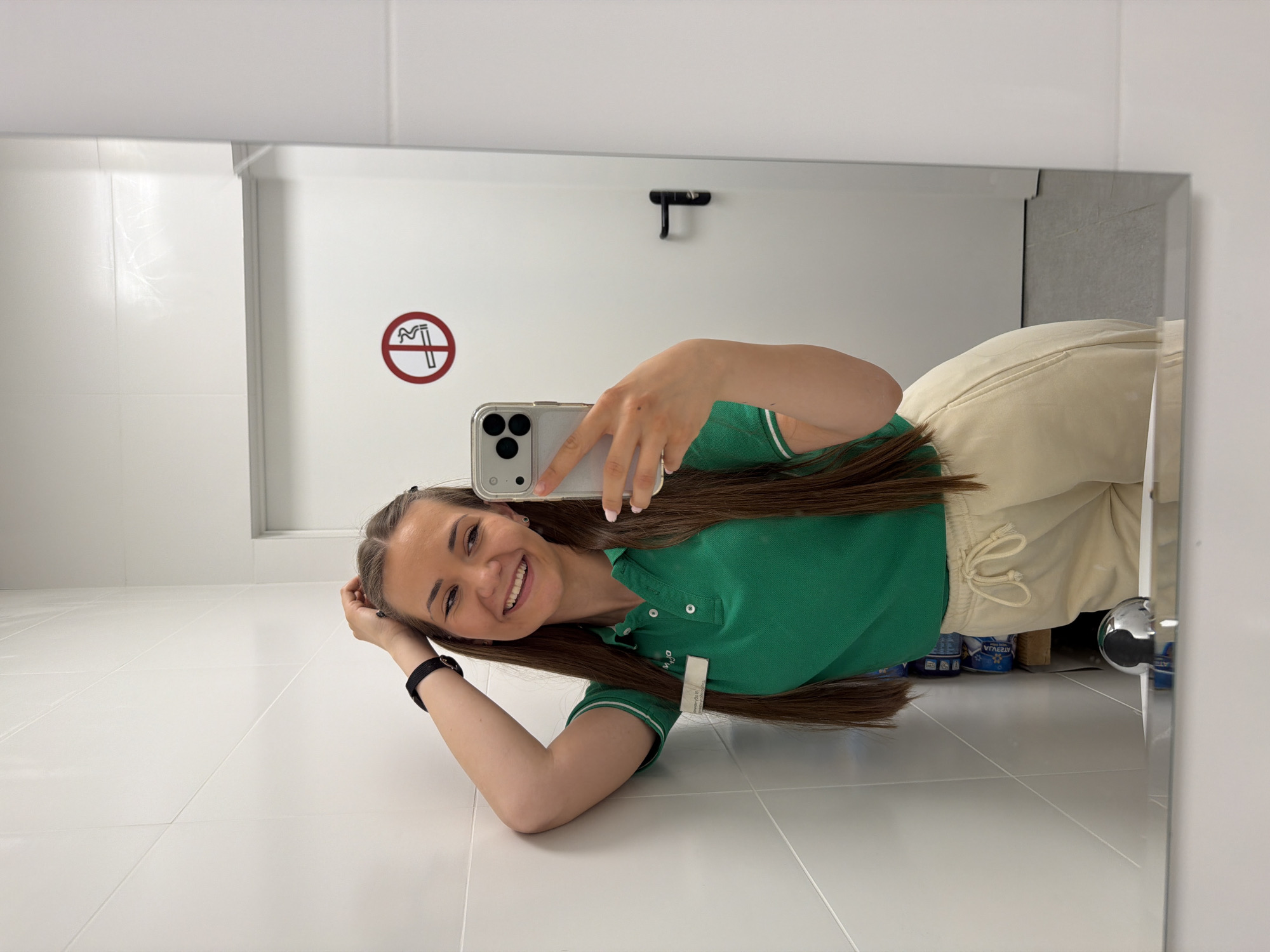 A young woman with long brown hair is taking a selfie in a mirror. She is wearing a green polo shirt with white trim and light-colored sweatpants. Her hair is down. She is smiling. She is holding a cell phone in her right hand. The mirror reflects a white tiled floor and wall with a no smoking sign. The location is likely a bathroom. The image provides no information to suggest the image was taken in the city of Sofia, Bulgaria.