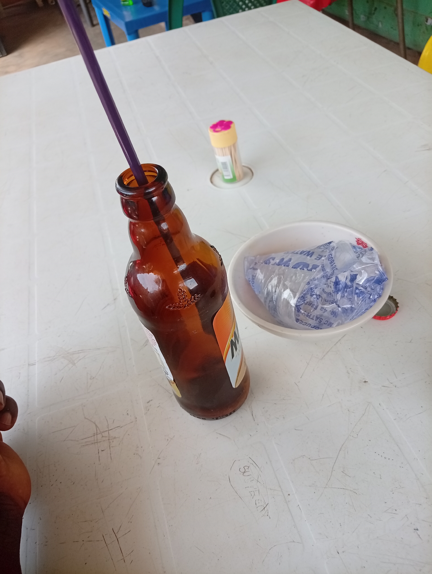 The image shows a slightly worn, light-colored plastic table surface with objects arranged on it. Dominating the left side is an open brown beer bottle with a purple straw inside. To the right of the bottle is a small white bowl containing a clear plastic bag filled with a clear, potentially wet, substance. Next to the bowl is a red bottle cap. Further back on the table, in the top center, is a small container of toothpicks. A small portion of a human hand is visible in the lower left corner of the image. The image was likely taken in Jos, Nigeria based on the style of objects and general environment.