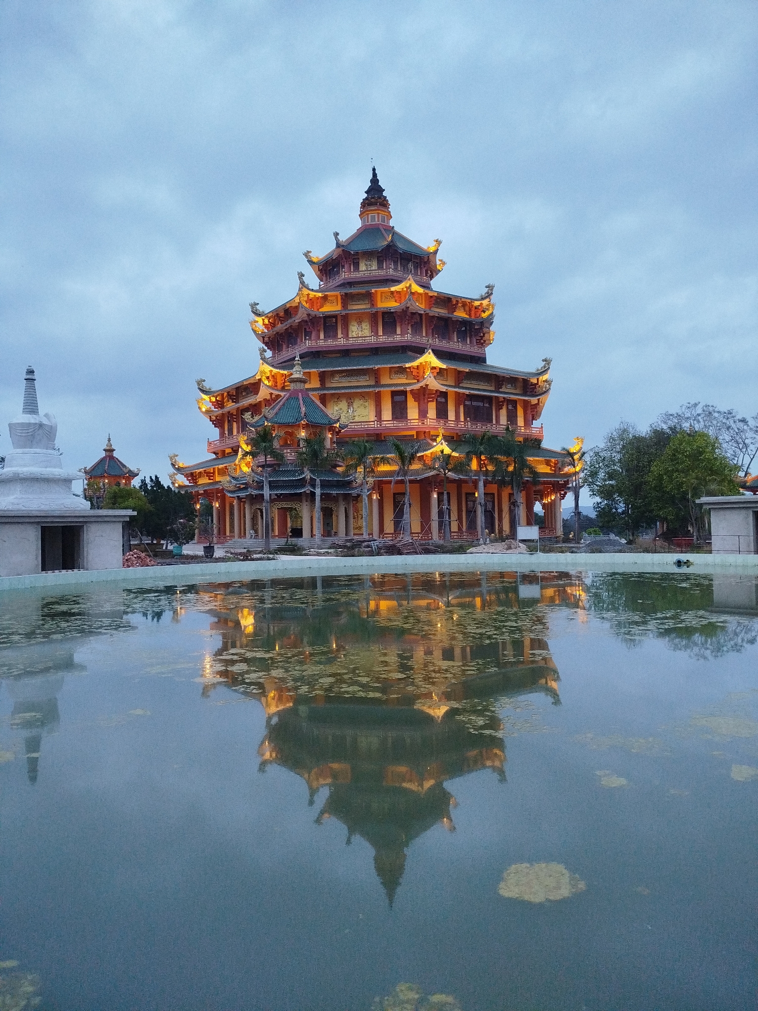 The image depicts a large, multi-tiered pagoda in Phú An, Vietnam, at dusk. The pagoda, illuminated with yellow lights, is reflected in a still body of water in the foreground. The structure is predominantly red and orange with green accents on the roof. The surrounding environment includes a white stupa-like structure to the left, other trees, and buildings in the background. The sky is overcast, creating a soft, diffused light.