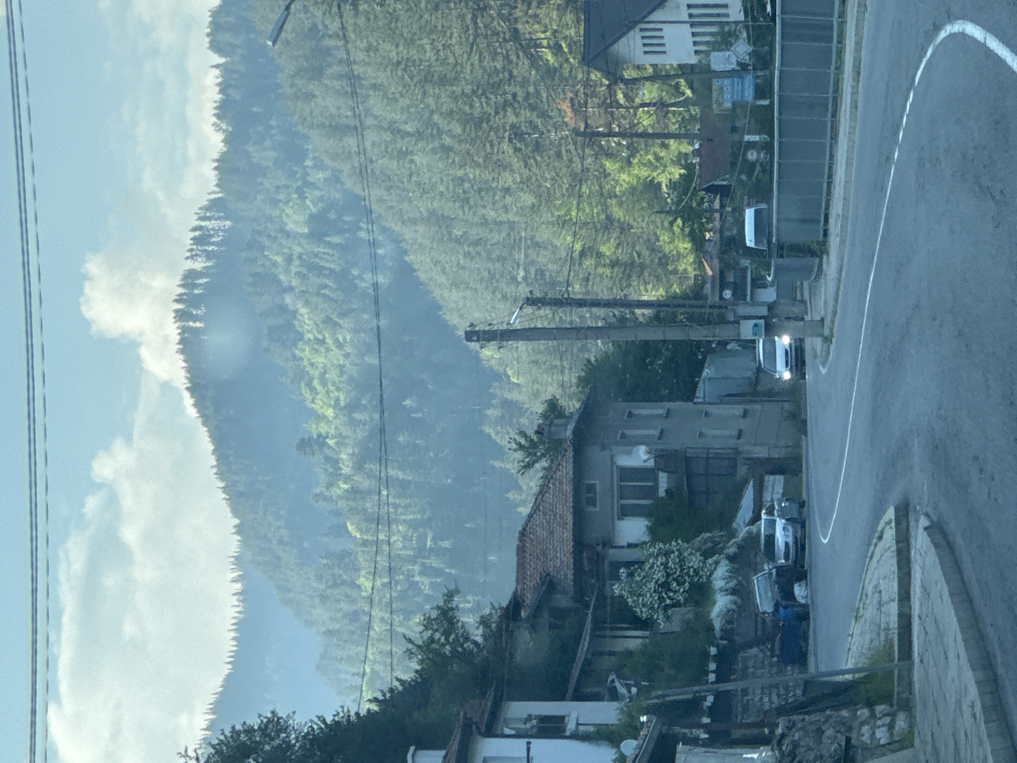 The image depicts a rural mountainous scene in Sokolovtsi, Bulgaria, likely during daytime under a partially cloudy sky with atmospheric haze. In the background, multiple layers of steep, densely forested mountains covered predominantly with coniferous trees rise under a pale blue sky. The distant mountain ranges appear lighter and less distinct due to the atmospheric haze.

In the midground, a paved road with a single white dividing line curves from the lower left towards the center and then right, continuing out of frame. On the left side of the road, several multi-story buildings are constructed into the hillside. These buildings feature traditional architecture with visible red-tiled roofs and light-colored (white or grey) facades. Dense green foliage, including trees and bushes with white blossoms, surrounds these structures. Two cars are parked on the left side of the road: a dark blue vehicle and a lighter-colored (possibly silver or white) sedan.

On the right side of the road, a metal barrier or railing runs parallel to the pavement. Further down the road on the right, a white building with a dark roof and multiple vertical window elements is visible. A white van or similar utility vehicle is parked near this building. A light-colored car with illuminated headlights is visible on the road, having just rounded a curve from the right.

Throughout the scene, multiple utility poles made of concrete or wood stand alongside the road, supporting a network of overhead power lines that traverse the sky and the landscape. A single street light fixture is visible on the upper right side of the image. No people are visible in the scene. The overall lighting suggests either early morning or late afternoon, characterized by a soft, diffused light and long shadows where visible.