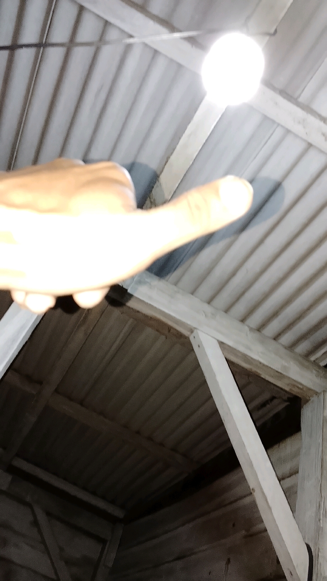 An overexposed, upward-facing, close-up shot shows a hand with its thumb extended, pointing towards a bright light bulb hanging from a wire, casting a shadow on a corrugated metal ceiling. The ceiling is supported by wooden beams, one of which is prominently displayed diagonally across the frame. Below the ceiling, wooden planks form a darker, unfinished structure. The scene appears to be an indoor space, possibly a shed or an unfinished building, located in Bojong Barat, Indonesia, given the context. The lighting is harsh and diffused, suggesting a low-light environment illuminated by a single, intense light source.