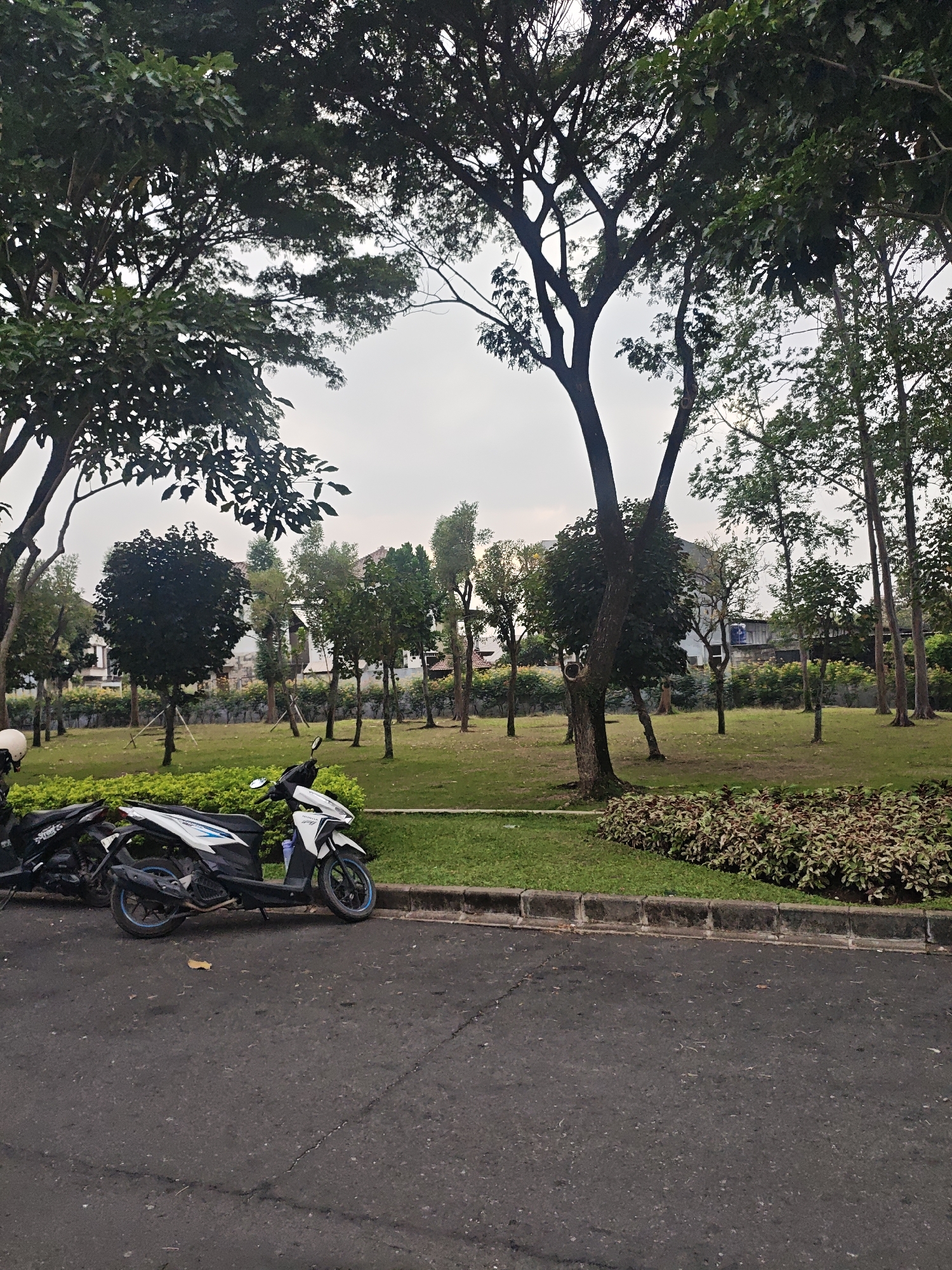 In the city of Bacin, Indonesia, a serene park scene unfolds under a hazy sky. Two parked motorcycles, one white and one black, rest on the asphalt edge of a road. Beyond the road, a lush green lawn is meticulously maintained, dotted with various trees. In the background, modern residential buildings with tiled roofs are visible, nestled amongst the foliage. The park features a diverse array of trees, from broad-leafed varieties to tall, slender ones, with some younger trees showing signs of recent planting. A decorative bed of red and green plants adds a touch of color to the landscape. The overall impression is one of a well-kept, modern residential area that incorporates green spaces for recreation and relaxation.