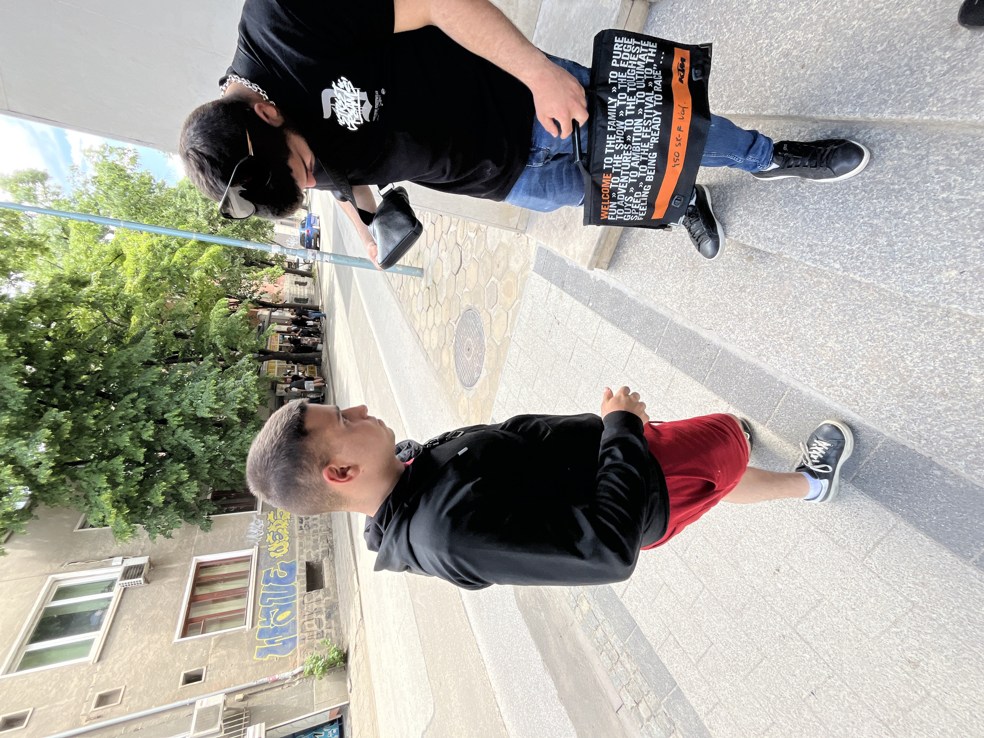 The image captures an outdoor street scene in Plovdiv, Bulgaria, on a bright, sunny day. Two young men are present on a paved sidewalk.
In the foreground, a young man with short, dark hair stands facing left, away from the camera. He wears a black hoodie, red shorts, and black sneakers with white socks. His hands are lightly clasped in front of him.
Behind him, closer to a building, is another young man seen from the side, partially obscured. He has dark hair with sunglasses resting on his head and wears a silver chain, a black t-shirt with a white graphic print, and blue jeans. He also wears black sneakers. He carries a small black shoulder bag and a large black and orange reusable bag in his left hand, while looking downwards.
The large black and orange bag features prominent text: "WELCOME TO THE FAMILY » TO PURE FUN » TO THE SHOW » TO THE EDGE GUYS » TO ADVENTURES » TO THE TOUGHEST SPEED » TO AMBITION » TO ULTIMATE THE FESTIVAL » THE FEELING BEING "READY TO RACE"." An orange stripe on the bag displays the KTM logo and "450 SK-F V.O.F.".
The ground is paved with light grey hexagonal and rectangular patterns. The background features lush green trees under a bright sky with some visible clouds. To the left, a light-colored building has multiple windows and a section of blue and yellow graffiti. The scene depicts a casual moment in an urban setting during what appears to be spring or summer.