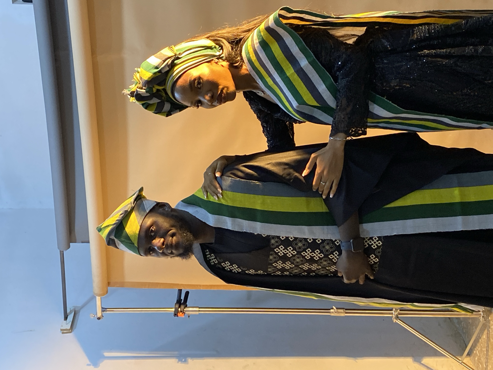 In Jos, Nigeria, a man and a woman stand in front of a backdrop. The man, facing left, wears a black outfit with a patterned vest and a traditional cap and sash with green, yellow, and white stripes. The woman, facing right, is dressed in a black sequined dress and a matching headwrap and sash. Her hand rests on the man's chest, and his hand is on his hip. A studio lighting stand is visible to the left of the frame.