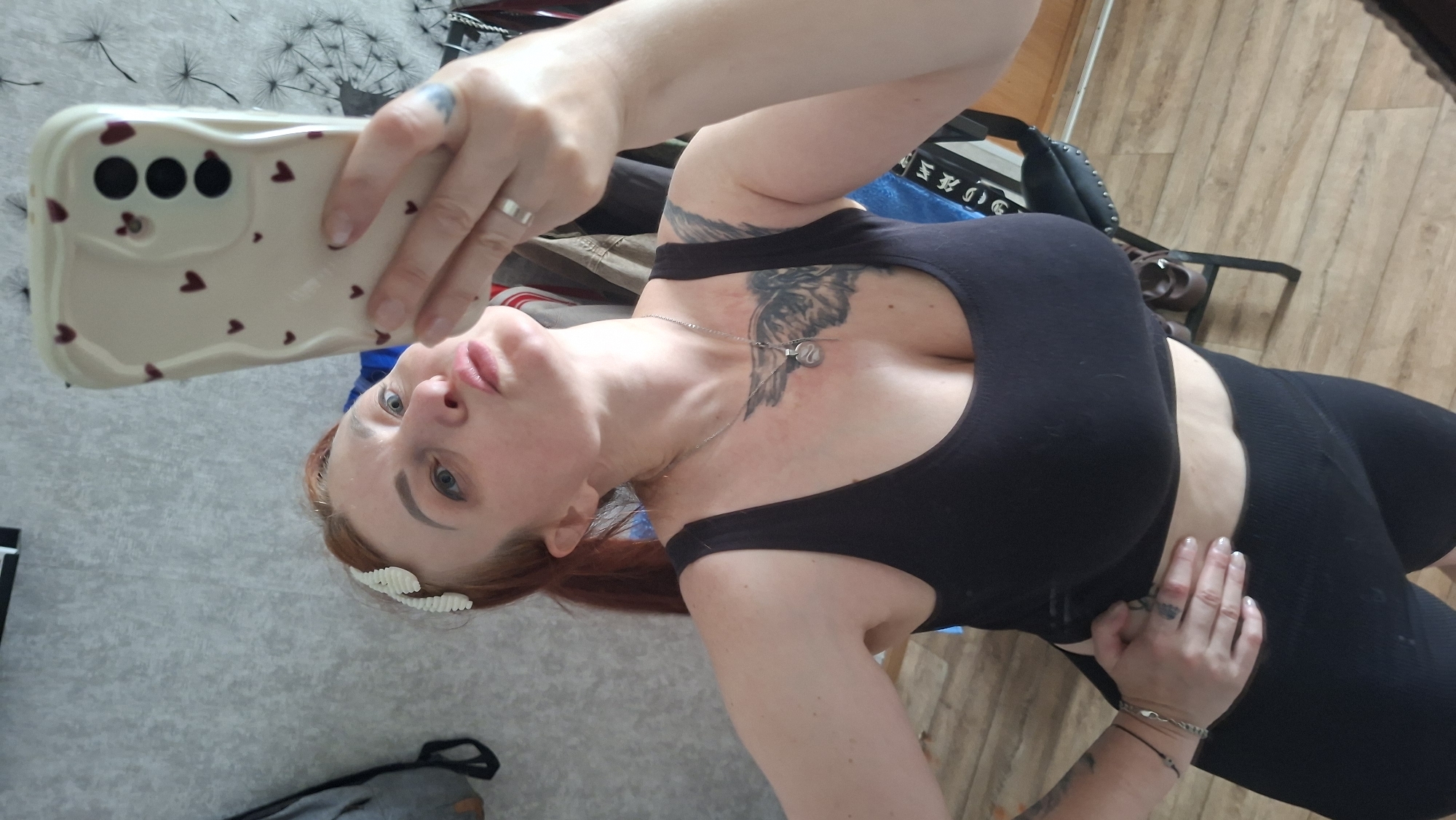 A woman with red hair pulled back in a ponytail and wearing a white hair clip is taking a selfie in a room. She is wearing a black sports bra and black shorts. She has a tattoo on her chest and arm. The background shows a grey wall and wooden floor. The woman is holding a white phone with pink hearts on the case. There is a chair and some clothing in the background.