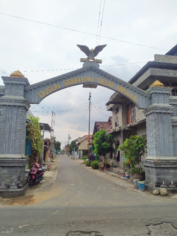 The image captures a static view of the entrance to a village or hamlet in Kecamatan Kebakkramat, Indonesia, characterized by a prominent decorative archway spanning a paved road. The archway, constructed from what appears to be grey concrete or stone with sculpted details, features golden-colored lettering on its curved beam that reads "DUSUN SOBRAYAN DESA BRUJUL". A large, dark-colored statue of an eagle with outstretched wings and a crest in its claws is mounted at the apex of the archway. The archway is supported by two robust pillars, each adorned with a golden, shell-like ornament on top.
The paved road extends straight into the distance, flanked by a mix of single and two-story residential buildings on both sides. These houses exhibit varying architectural styles, some with exposed concrete and others with tiled roofs. Along the sides of the road, particularly in front of the houses, there are various potted plants and small trees, adding greenery to the scene. On the left side of the road, just past the archway, a red scooter is parked. Utility poles with numerous overhead wires are visible along the road, indicating a typical village infrastructure. The sky is overcast with a blanket of grey clouds, suggesting either an early morning, late afternoon, or simply a cloudy day. The overall impression is that of a quiet, rural or semi-urban setting. No people are visible, and no specific activities are taking place.