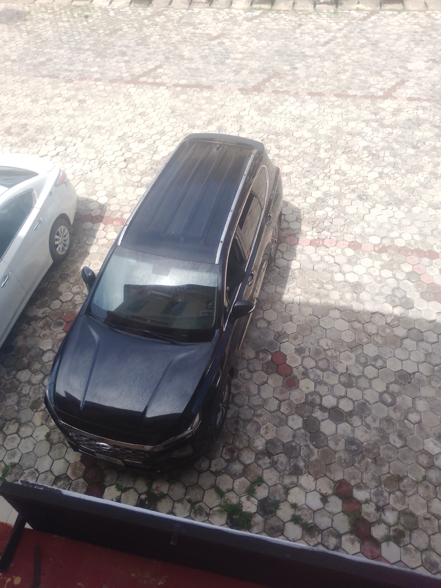 In Gura Top, Nigeria, a dark-colored SUV is parked on a hexagonal-tiled surface. The vehicle, seen from a high angle, is oriented diagonally. Adjacent to the SUV, a white sedan is also parked. The ground is a textured pavement with visible seams between the hexagonal tiles. Shadows suggest sunlight casting onto the scene from the right. There is some sparse vegetation growing between the tiles. The image captures an outdoor parking area.