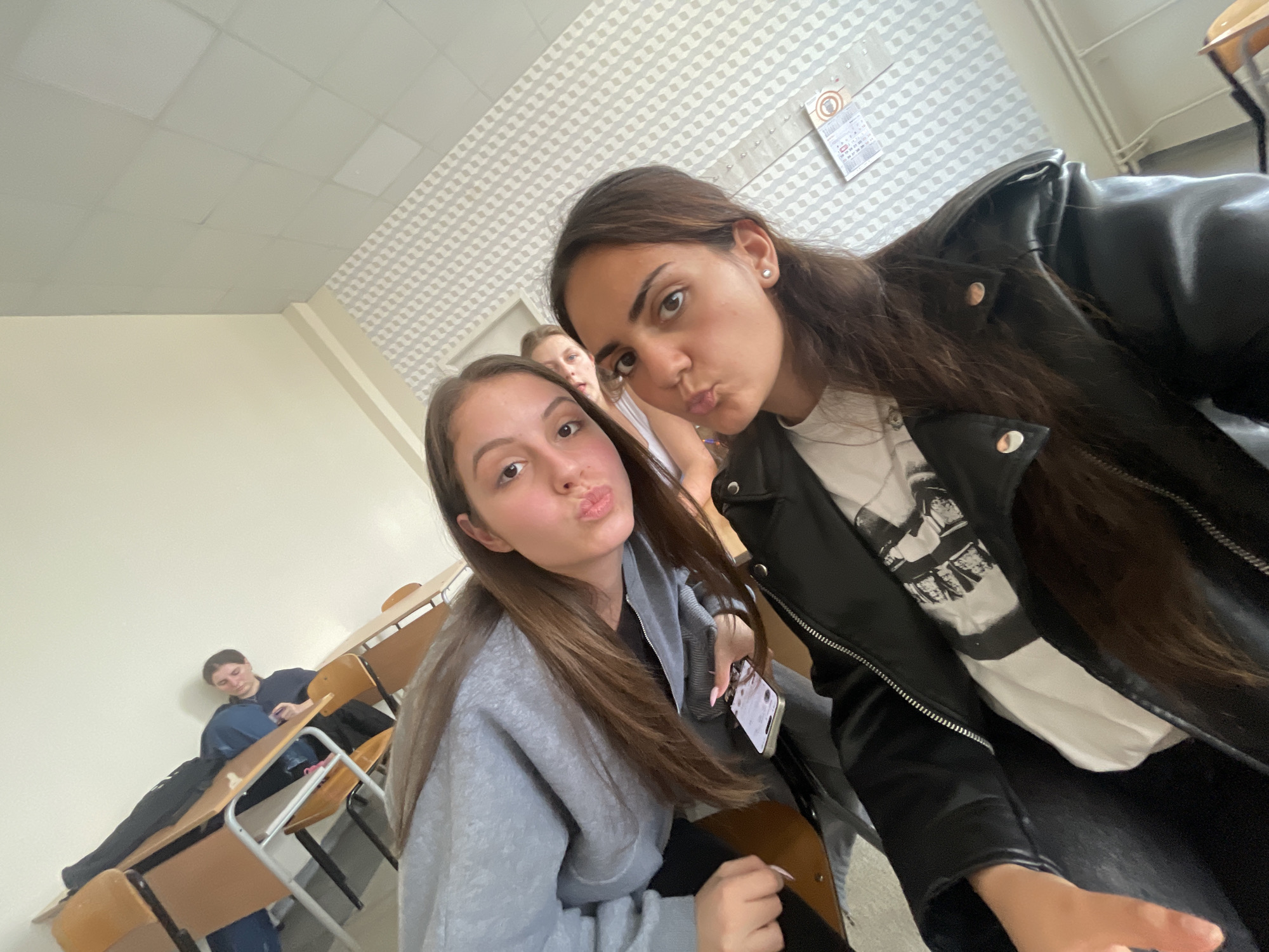 The image appears to be taken indoors, in what looks like a classroom setting. Three young women are the primary focus, with two of them positioned in the foreground striking a pose with puckered lips, as if taking a selfie. The woman on the right is wearing a black leather jacket over a white t-shirt, while the one on the left is wearing a gray sweatshirt. The third woman is partially visible in the background between the two women in the foreground. A person is sitting at a desk at the background. Rows of empty desks and chairs are visible. The ceiling has a tiled pattern, and a calendar is affixed to the wall. The overall lighting is bright and even. The image is angled, suggesting it was taken with a handheld device. There is nothing to suggest this image was taken in the city of Burgas, Bulgaria.