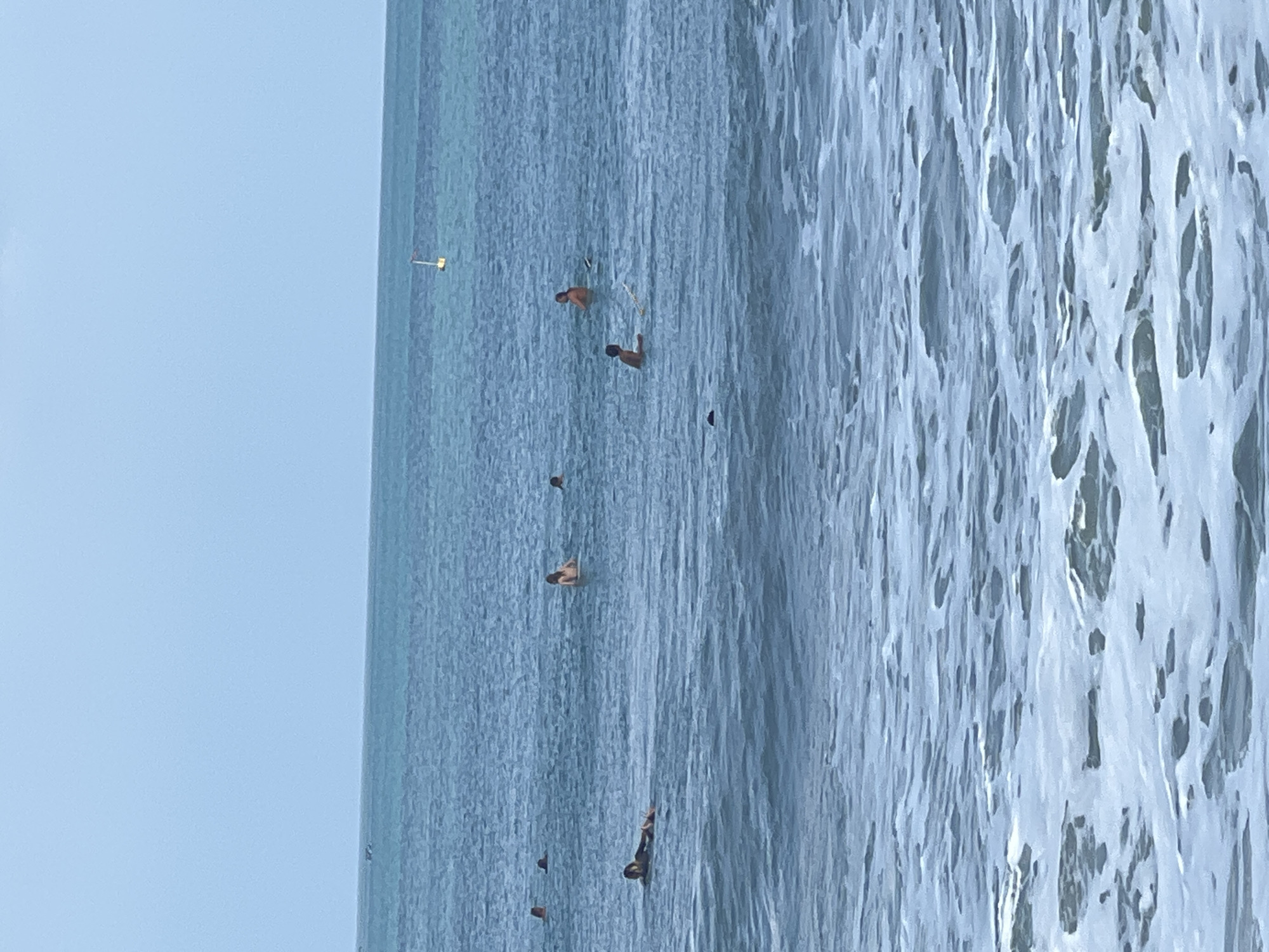 This image shows a view of the ocean in Kuta, Indonesia, with several people in the water. The water is blue with some white foam from waves in the foreground. In the midground, there are at least seven individuals, some appearing to be surfing or paddling on surfboards, and others just in the water. A yellow buoy with a red top is visible in the distance. The sky is a pale blue.
