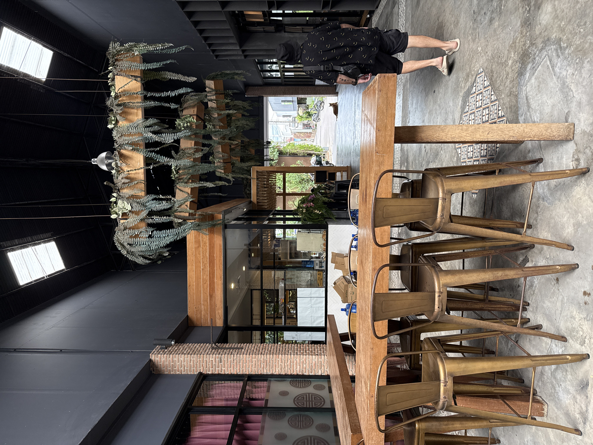 A man wearing a black bucket hat, a black short-sleeved shirt with a smiley face pattern, and black shorts walks away from the camera towards an open pathway. He has a black crossbody bag slung over his right shoulder and is wearing light-colored slide sandals. In the foreground, a long wooden table is set with several metallic gold bar stools. The scene appears to be an outdoor dining or seating area. Behind the table, to the left, is a building with large windows and brick accents, draped with pink curtains. Hanging plants are suspended from wooden beams above. The pathway leads to a street with buildings and parked motorcycles. A cat is lying on the ground in the pathway. The overall environment suggests a commercial establishment in a tropical location, possibly a cafe or restaurant in Ko Samui District, Thailand.