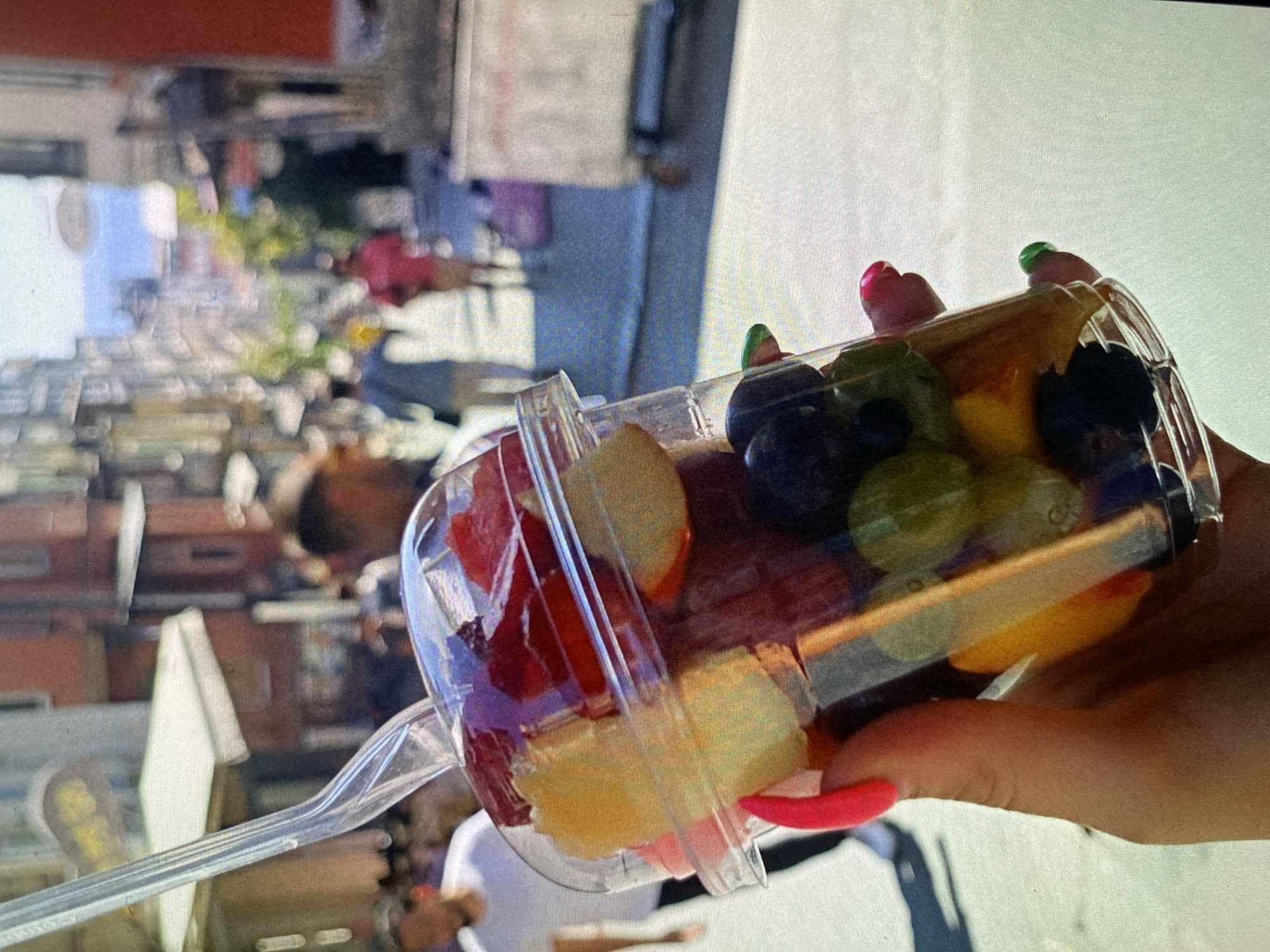 The image shows a close-up of a person's hand holding a clear plastic cup filled with various chopped fruits. The fruit assortment includes red pieces (possibly strawberries or watermelon), green grapes, dark blueberries, and lighter yellow/white pieces (likely pineapple, melon, or apple). A clear plastic spork is inserted into the cup. The hand has long fingernails painted in bright colors; most are a vivid pink or red, while one nail is green.
The background is significantly blurred, indicating a shallow depth of field, which keeps the fruit cup as the primary subject in focus. Despite the blur, the setting appears to be an outdoor urban or semi-urban environment during daytime. Outlines of buildings and indistinct figures of people are visible in the background, suggesting a bustling public area like a street, market, or park. The bright lighting indicates a sunny day. The activity captured is a person enjoying a fresh fruit snack, likely on the go or while relaxing outdoors. No visible text is legible in the image. Based on the provided context, the scene is located in Bankya, Bulgaria.