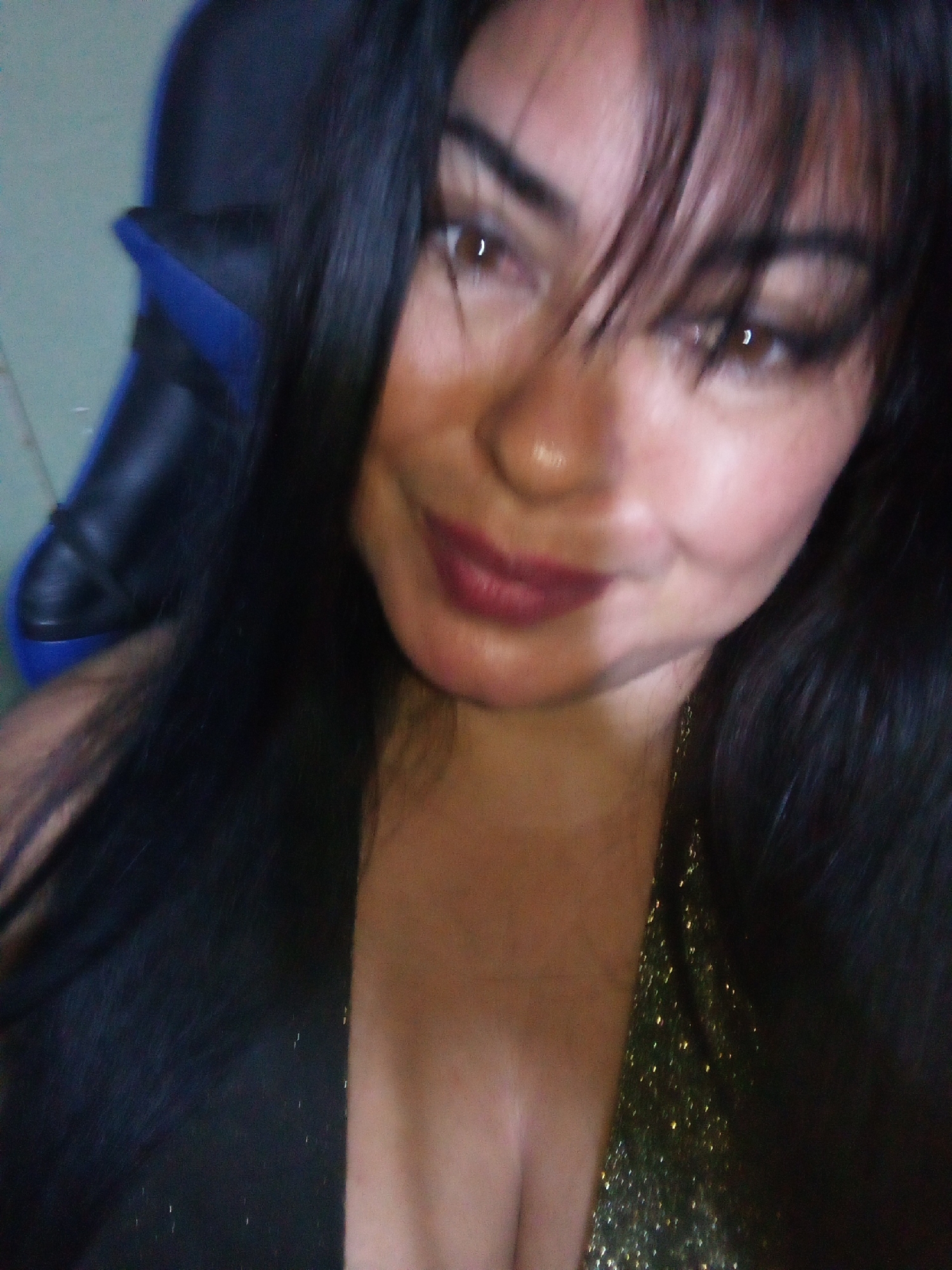 A close-up photograph depicts a woman with dark hair, styled with bangs. She has brown eyes and wears dark lipstick. The background shows a blue and black chair, and a glimpse of a light green wall. The woman's clothing appears to be dark, possibly a dress, with some gold sparkly accents visible. The picture's location in Juan Griego, Venezuela cannot be ascertained from the provided details.