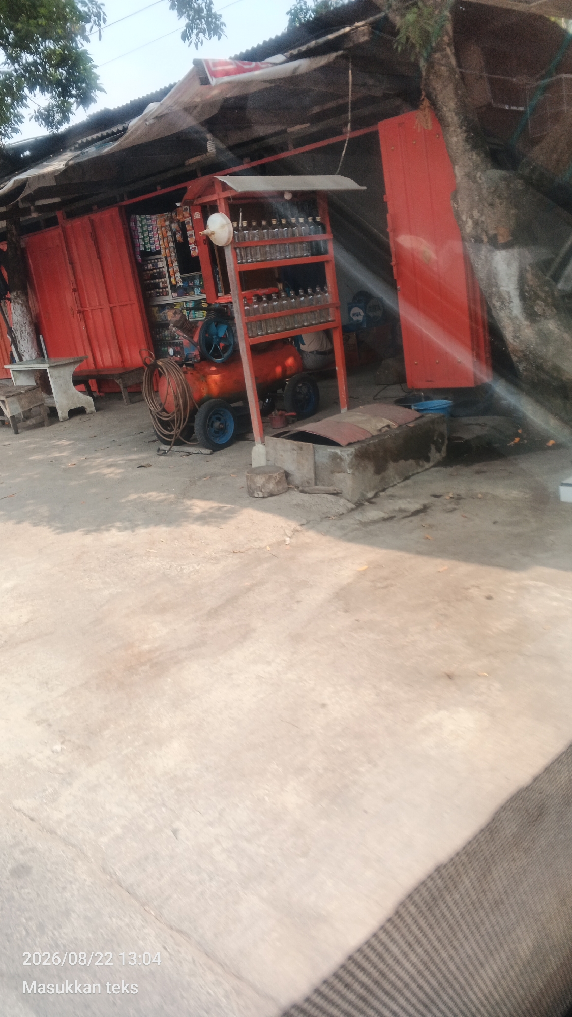 The image shows an outdoor scene in Sukoanyar, Indonesia, featuring a small, open-air store or stall. The stall has a red metal structure, with shelves displaying various bottles, possibly for sale. A large orange air compressor with a coiled hose and blue wheels is visible in front of the stall. Adjacent to the stall, a tree trunk grows near the structure. The ground is paved with concrete and shows signs of wear and tear. In the foreground, a portion of a vehicle's interior, possibly a car, is visible, indicating the photo was taken from inside a vehicle. The overall impression is that of a typical roadside vendor setup in a tropical environment. The date and time stamp at the bottom left corner reads "2026/08/22 13:04".