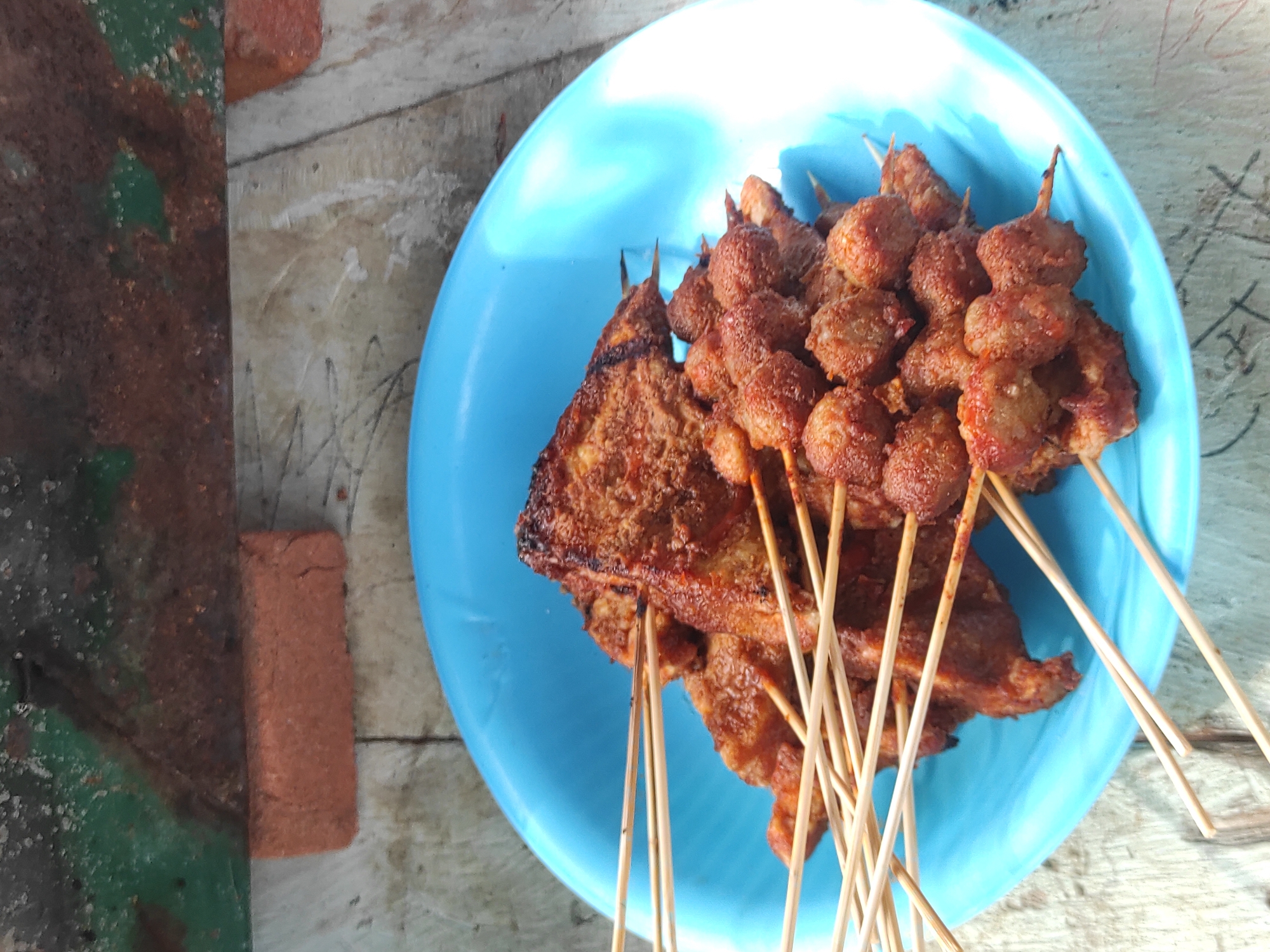 The image captures a slightly elevated, close-up perspective of a blue plastic bowl filled with numerous wooden skewers of cooked food, situated on a textured, light-colored surface. In the background, a single reddish-orange brick rests horizontally, backed by a dark, mottled surface that displays patches of green and substantial reddish-brown coloration consistent with rust or heavily weathered material.
The blue plastic bowl contains two visually distinct types of skewered food. One type consists of flattened, somewhat irregular or triangular pieces, heavily coated in a coarse, dark reddish-brown seasoning or sauce. The other type features clusters of small, roughly spherical or nugget-like pieces, similarly coated in a reddish-brown substance. Both food types exhibit dark, caramelized, or possibly charred areas, indicative of grilling or frying. Multiple thin, light-colored wooden skewers are arranged within the bowl, some protruding prominently.
The immediate surface supporting the bowl is a light, off-white or beige material, possibly concrete or a worn table, marked with visible lines, subtle textures, and some faint dark smudges or scuffs. The scene is illuminated by bright, natural light, creating some reflections on the blue bowl. There are no individuals visible in the frame. The setting, as specified, is within the city of Lhokseumawe, Indonesia.