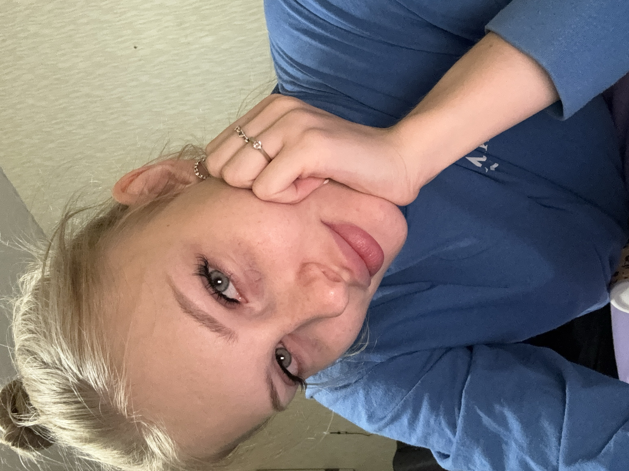 A young woman with blonde hair styled in a bun is pictured in a close-up selfie. She is resting her head on her hand, which is adorned with two rings on her fingers. Her eyes are a striking blue-green, and she appears to be wearing subtle makeup, including mascara and lip gloss. She is wearing a blue long-sleeved shirt. The background is a plain, textured wall. The image does not provide any information about a specific location within the city of Kondratovo, Russia.