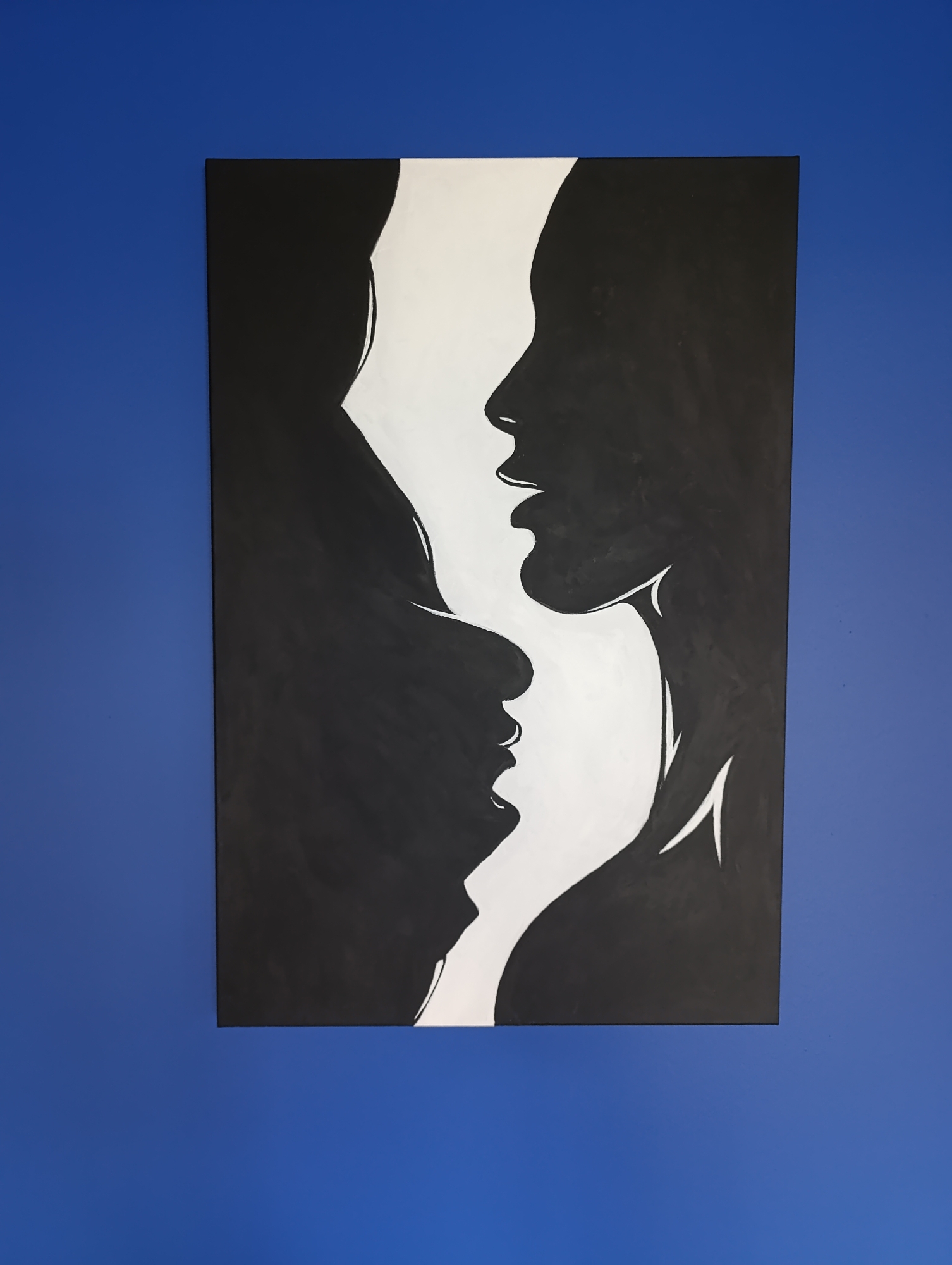 The image displays a black and white painting on a blue wall. The painting is abstract and features the silhouettes of two profiles facing each other, with a white space in between them. The profiles appear to be of human faces, one larger and positioned above the other. The larger profile's head is tilted downwards, while the smaller profile is looking upwards. The painting is mounted on a solid blue wall. There is no information available in the image to suggest a location in Minsk, Belarus.