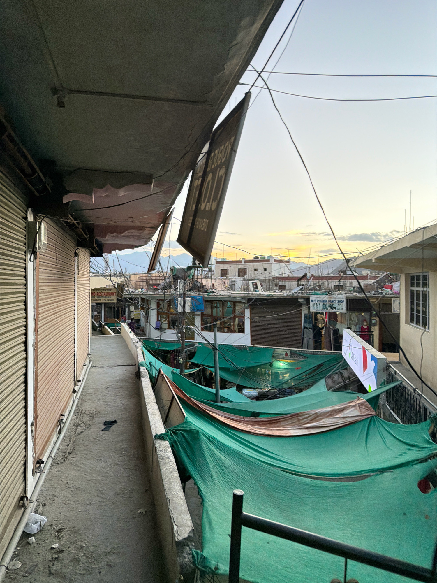 The image depicts a view of a street in Leh, India, likely in the evening with the sky displaying hues of orange and yellow from the setting sun. The perspective is from a raised walkway or balcony, looking down onto a street lined with shops and buildings.
On the left, a shop with a corrugated metal shutter is visible, its brown surface showing signs of wear. A pipe runs along the side of the building. The walkway itself is made of concrete and has a rough texture, with some debris and a plastic bag on the ground.
To the right of the walkway, there is a railing, and beyond that, several shops are visible. Many of these shops are covered with green tarpaulins or fabric awnings, suggesting they might be temporary stalls or market stalls. One of these tarpaulins has a tear. There are also some signs on the buildings, including one that reads "M S CHENBA FANCY SHOP".
Further in the background, other buildings with white facades and windows are visible, and mountains can be seen in the distance. There are numerous electrical wires crisscrossing the scene, adding to the cluttered urban environment. A few people are sparsely visible in the background, appearing to be shoppers or residents.
A black sign hangs from an overhead structure in the center of the image, tilted at an angle, with some text on it that is partially obscured and illegible from this distance. Another white sign with some colorful elements and text is also visible in the foreground on the right, partially obscured by the green tarpaulins. The overall impression is of a busy, somewhat unkempt, yet vibrant market street in Leh.