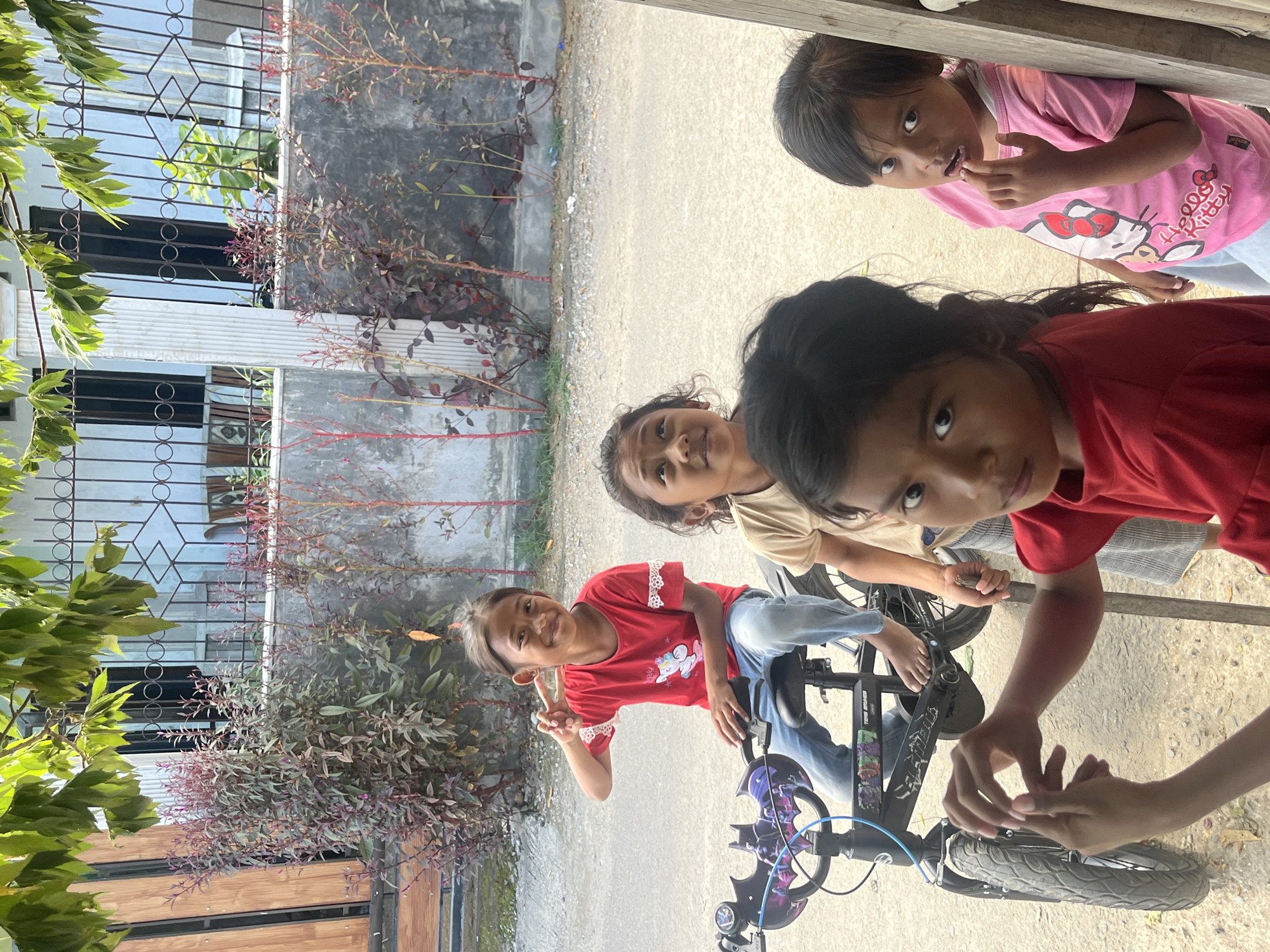 In Kertasari, Indonesia, four young girls are gathered outdoors near a street. A girl in a red shirt and blue jeans is sitting on a black bicycle, posing with a peace sign. To her right, another girl in a tan shirt looks upwards. In the foreground, a girl in a red shirt and a girl in a pink Hello Kitty shirt are looking towards the camera. The background features a fence, plants, and a building.