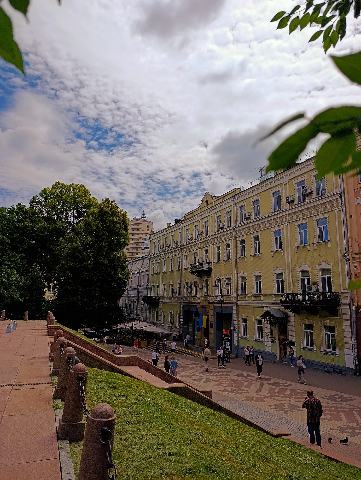 This image depicts a view of St. Andrew's Descent in Kyiv, Ukraine. The scene is dominated by a row of historical buildings lining the street, with a prominent yellow building in the foreground that features ornate white trim around its windows and balconies. The street itself is paved with a decorative pattern of red, beige, and grey tiles. Several people are visible walking along the street and sidewalks, some appearing to be tourists or locals engaged in daily activities. To the left of the street, a grassy embankment slopes upwards, bordered by a red stone staircase and a series of stone bollards connected by chains. Large trees with dense green foliage stand behind the buildings, and a taller, modern building is visible in the background. The sky above is filled with a mixture of white and grey clouds against a blue sky. In the lower right corner, a man in a plaid shirt is standing on the grass, looking at his phone. Two pigeons are on the ground near him.