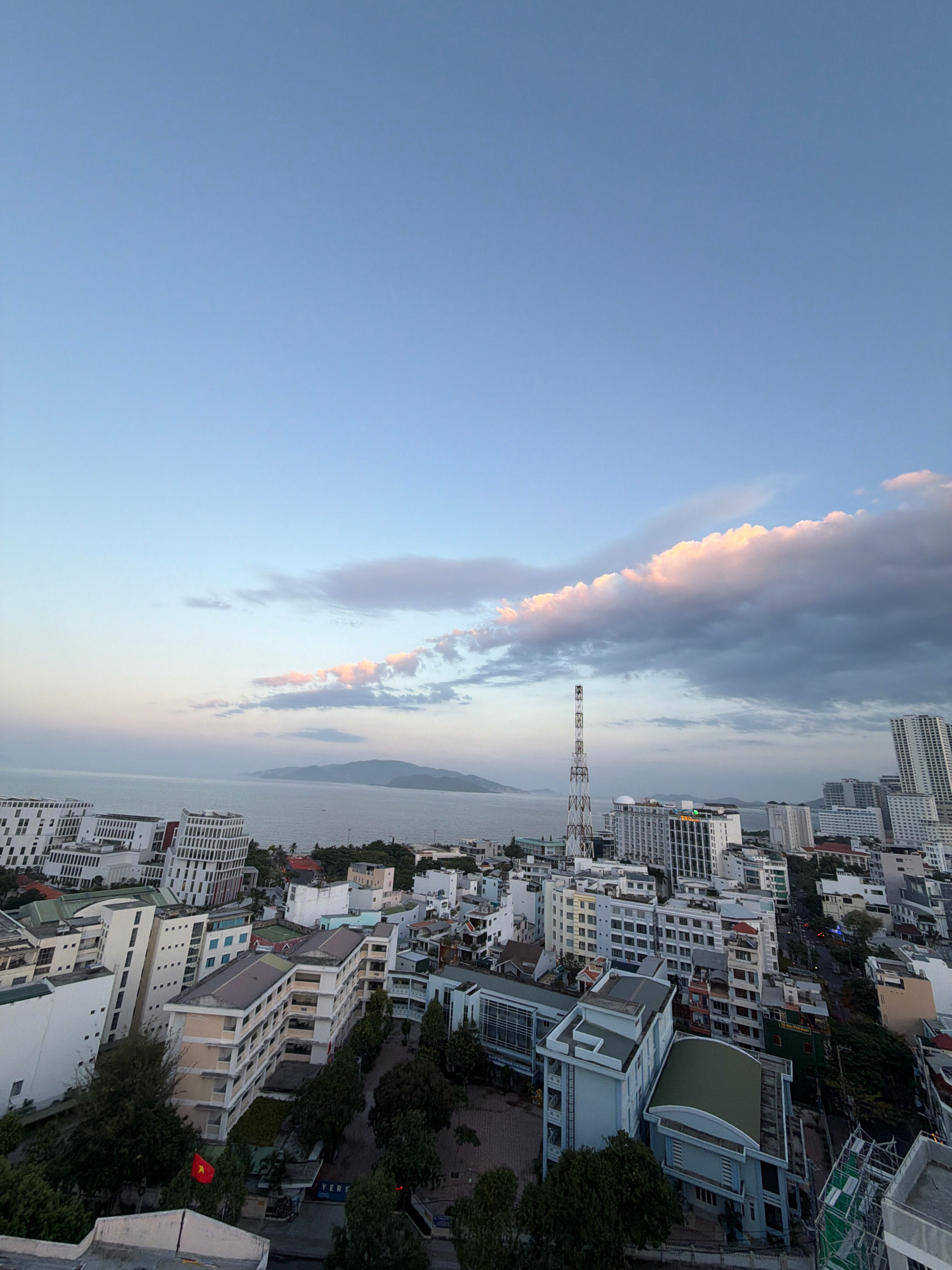 This is an aerial, wide-angle photograph of the coastal city of Nha Trang, Vietnam, taken from a high vantage point, possibly a tall building. The sky is a pale blue with scattered clouds tinged with pink and white, indicating either sunrise or sunset. In the distance, a hazy mountainous island is visible on the horizon, separated from the city by a body of water.
The foreground and midground are filled with densely packed buildings of varying heights and architectural styles. Many are white or light-colored, with flat roofs, and some exhibit modern designs with many windows. A prominent metal telecommunications tower stands tall among the buildings. Trees are scattered throughout the urban landscape, particularly in the lower right portion of the image, where they appear denser. A Vietnamese flag, red with a yellow star, is visible flying from a pole amidst the greenery in the lower left. The overall impression is of a bustling coastal city with a blend of modern and older structures, set against a natural backdrop of sea and mountains. There are no visible people or ongoing activities.