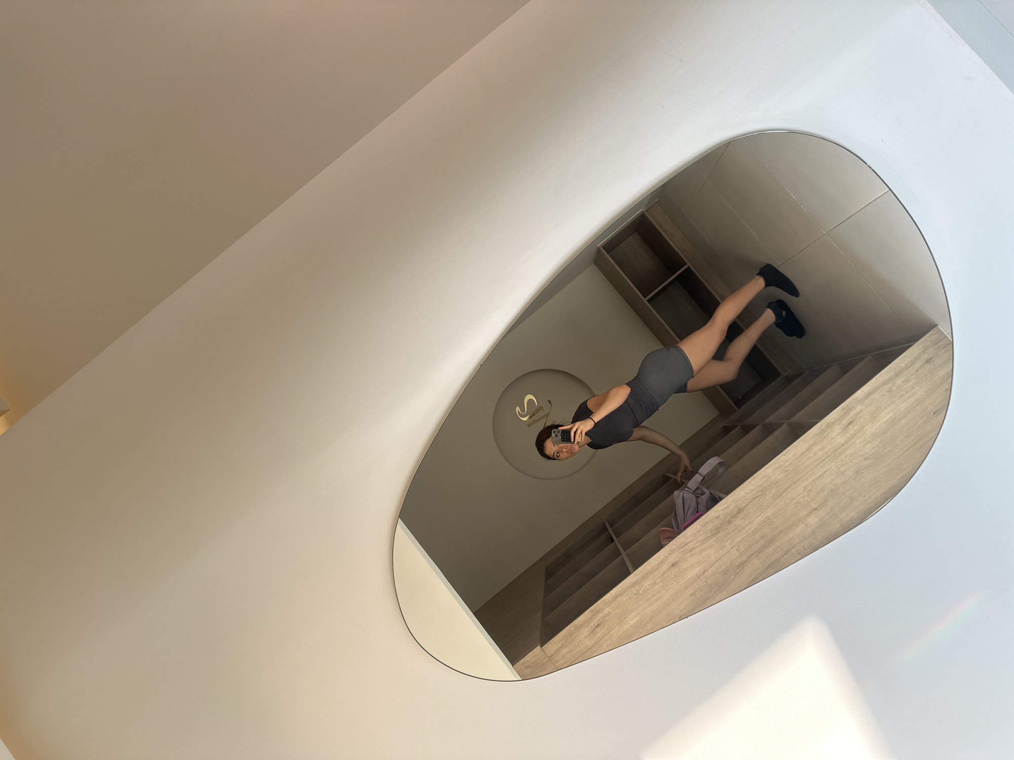 A woman in a black shirt and gray shorts takes a selfie in a large, oddly-shaped mirror. The mirror reflects a wall with wooden shelves. The background appears to be a room with a beige ceiling and wall. The location cannot be determined.